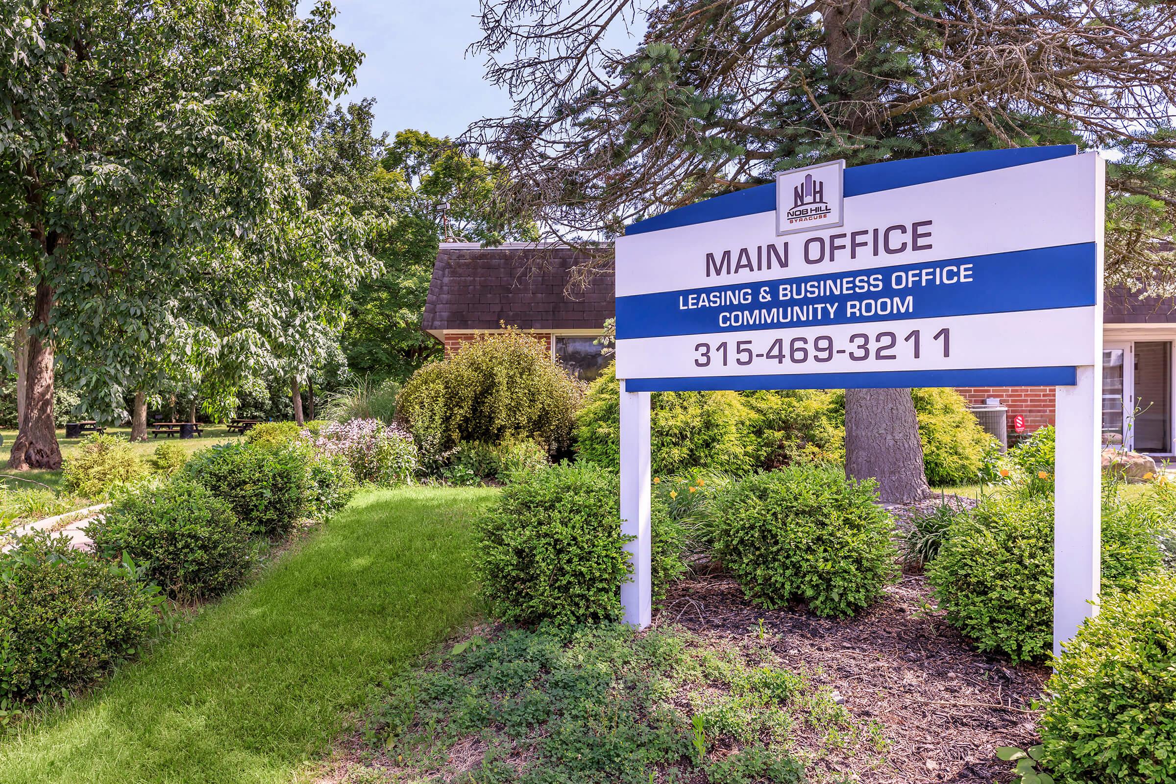 A well-maintained exterior view of a building with a sign that reads "MAIN OFFICE" along with "Leasing & Business Office" and "Community Room." The area features lush greenery, bushes, and trees, indicating a welcoming atmosphere.
