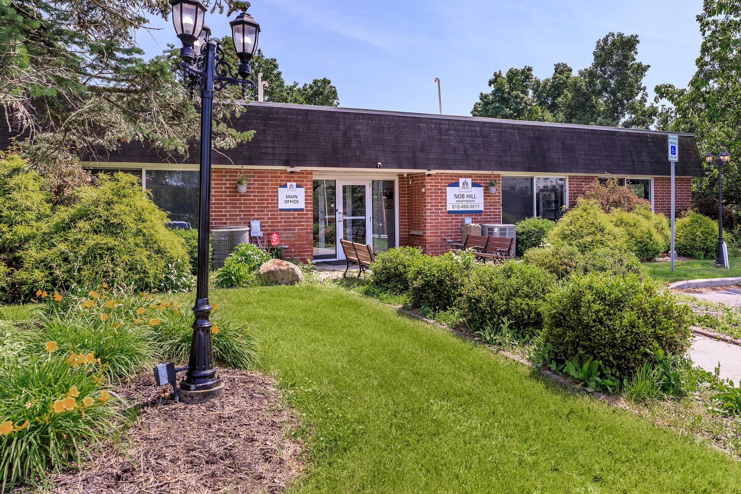 A view of a single-story brick building featuring large windows and signage that reads "Pool Office" and "203 Hill." The area is surrounded by well-maintained greenery, including bushes and flowers, with a lamp post in the foreground and clear blue skies above.