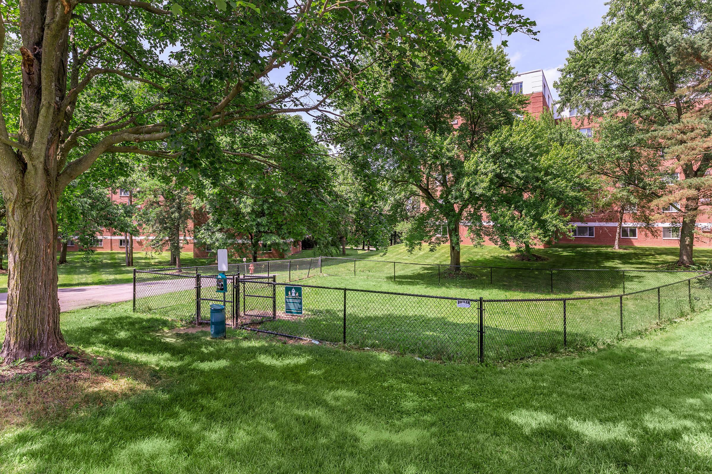 A green, grassy area enclosed by a black fence, featuring several trees providing shade. In the background, a building with a red brick facade is partially visible. A pathway runs alongside the area, indicating a well-maintained outdoor space.