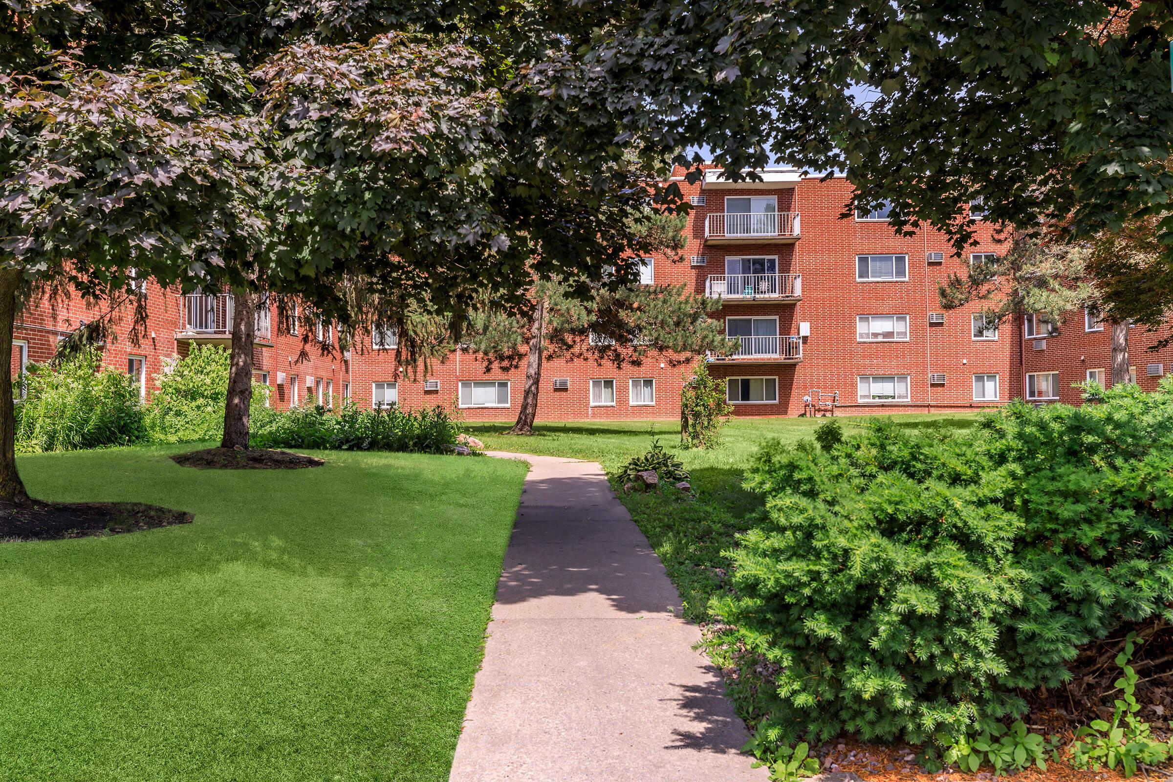 A pathway leads through a green lawn area surrounded by trees, with a view of a red brick apartment building in the background. The building features multiple balconies and windows, creating a pleasant residential environment.