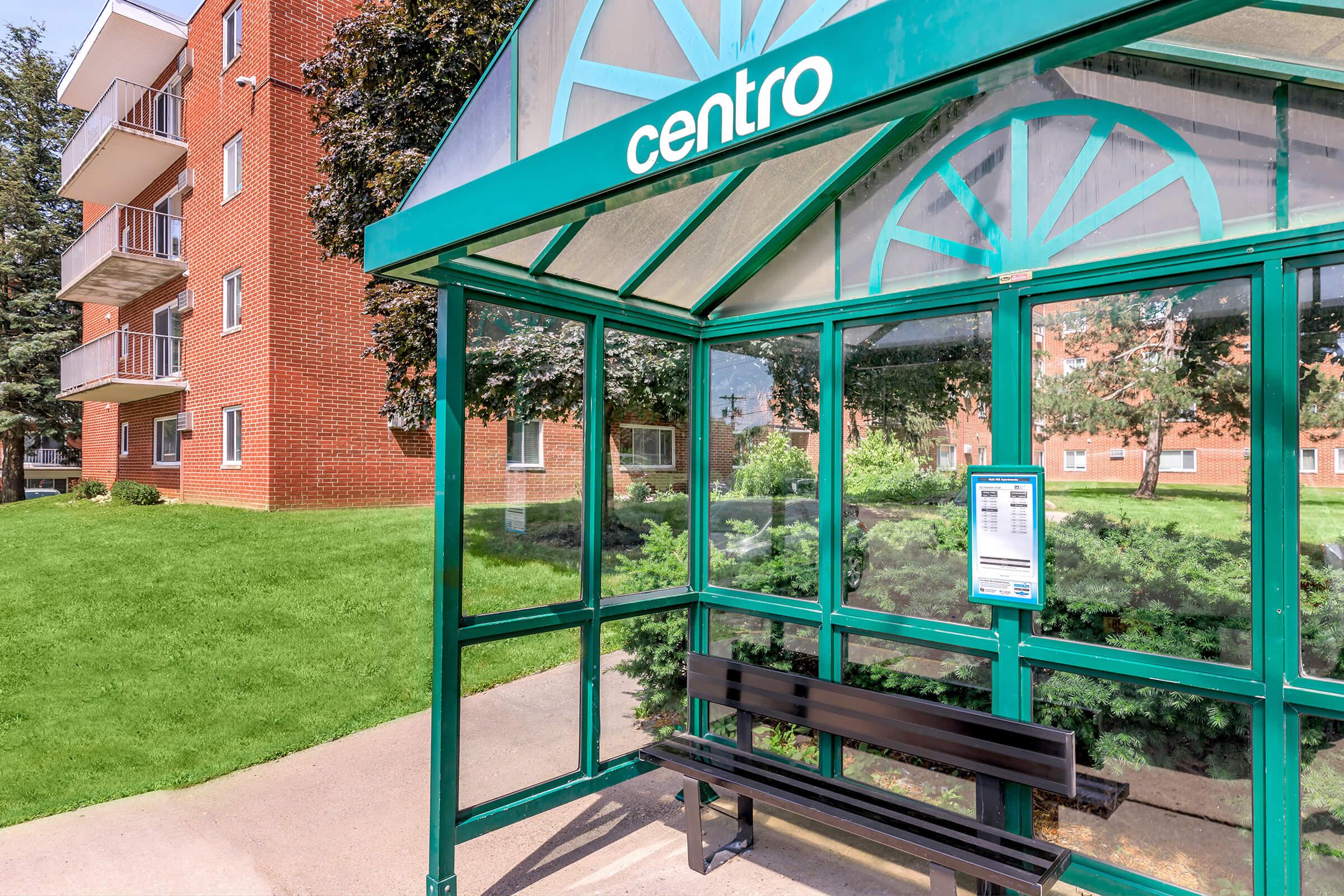 A bus shelter with a green frame labeled "centro" is shown. It features a seating bench and a schedule display on the side. In the background, there are brick apartment buildings and a well-maintained grassy area. The scene is bright and sunny, indicating good weather.