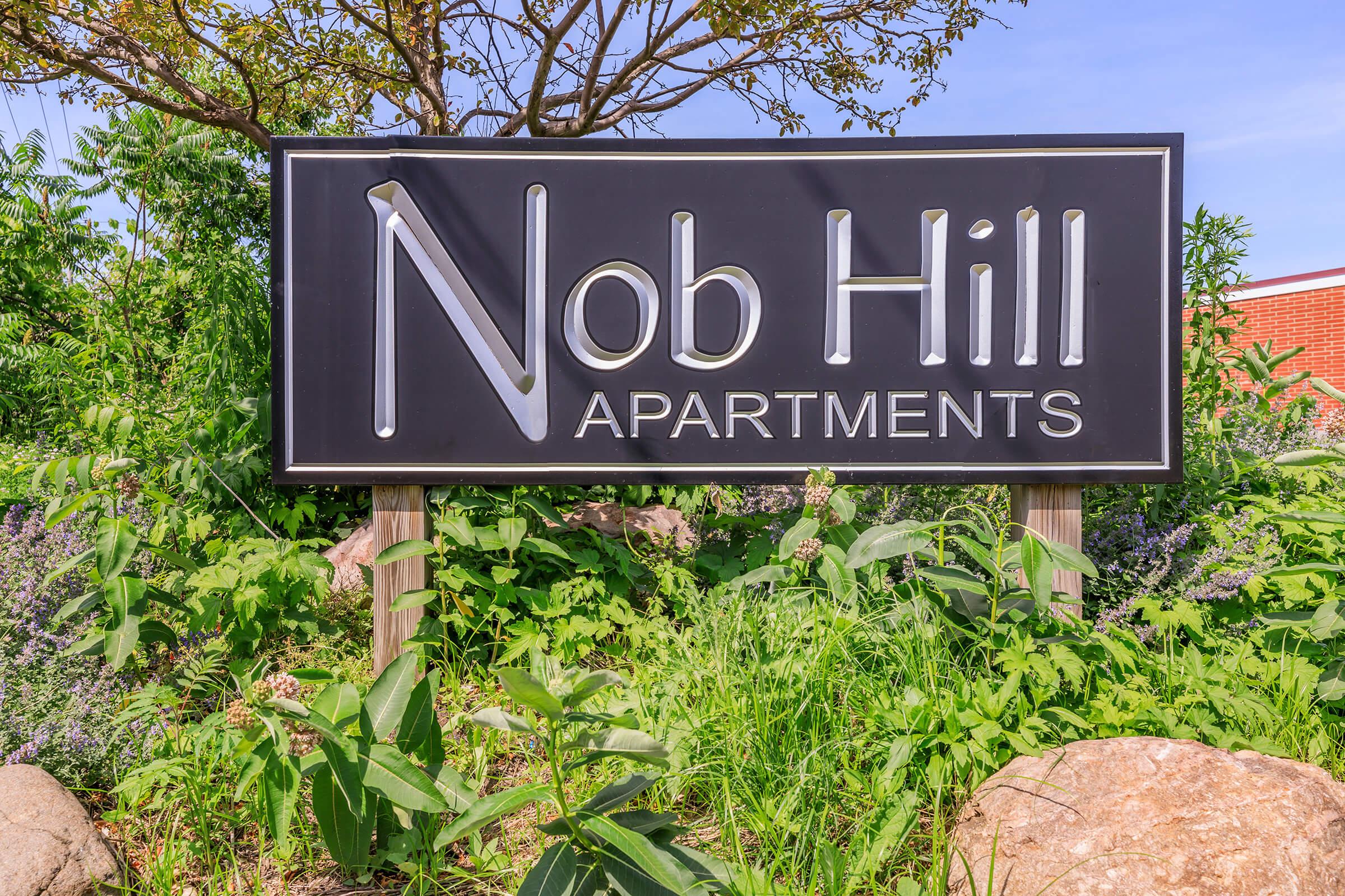 A sign for Nob Hill Apartments, featuring white lettering on a black background, surrounded by lush green foliage and flowers. The setting is bright and sunny, indicating a welcoming environment for residents.
