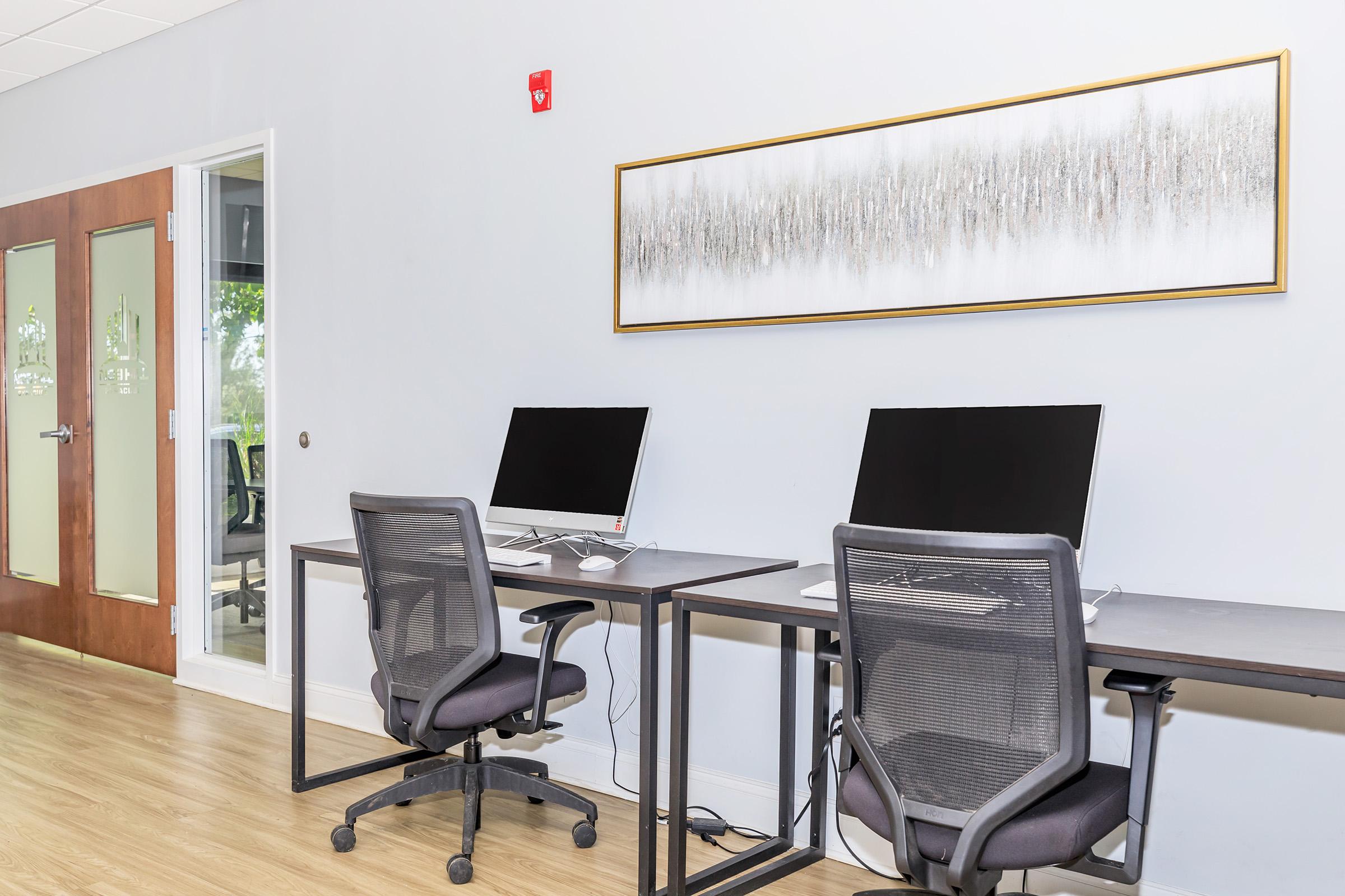 Two modern desks with black computer monitors and ergonomic chairs in a minimalist office space. The walls are painted light gray, and there's a decorative framed artwork depicting abstract trees. A glass door leads to another room, allowing natural light to brighten the area.