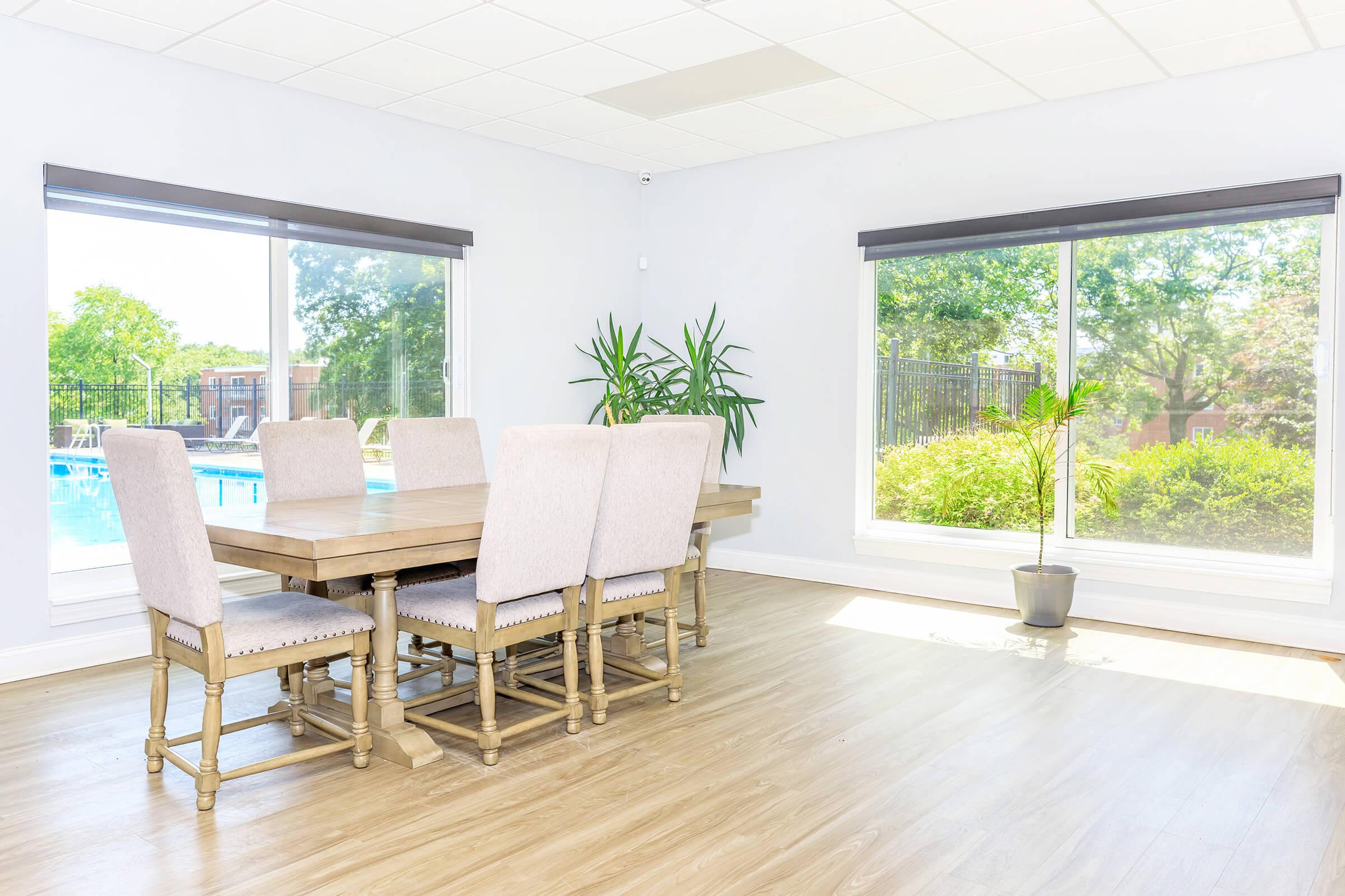 A bright and airy dining area featuring a wooden table with eight upholstered chairs. Large windows let in natural light, revealing a view of greenery and a swimming pool outside. Potted plants are placed in the corners, enhancing the open and welcoming atmosphere.