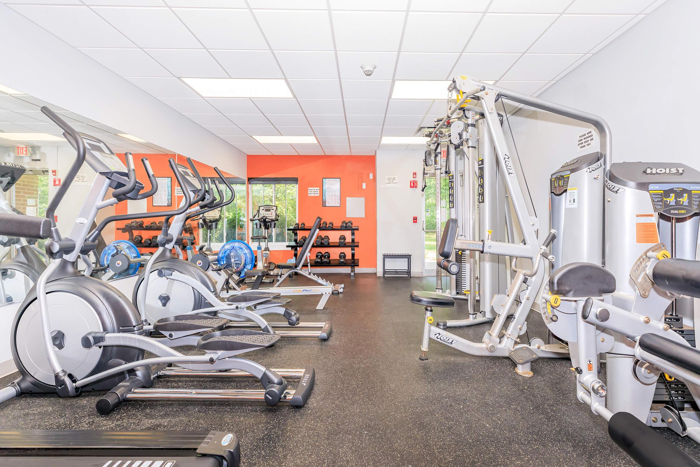 A modern fitness room featuring various exercise equipment including elliptical machines, a weight machine, and free weights. The room has bright orange walls, large mirrors, and a clean, organized layout, creating an inviting space for workouts.