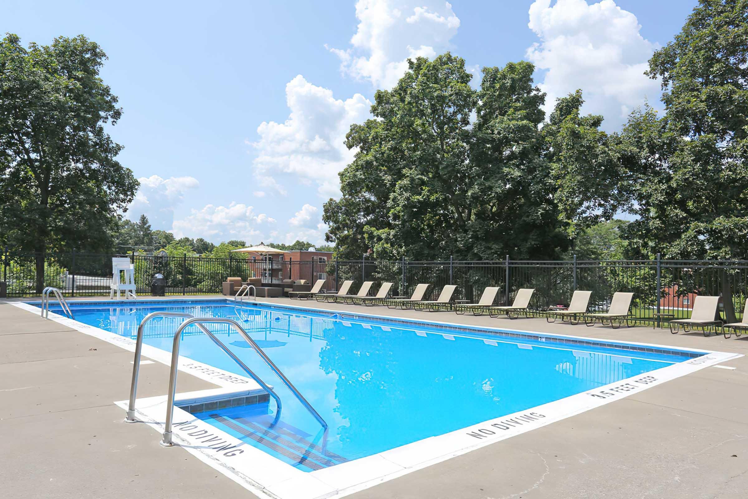 A clear blue swimming pool surrounded by lounge chairs on a sunny day. Lush green trees provide shade around the area, and a "No Diving" sign is visible near the pool's edge. The sky is mostly sunny with a few fluffy clouds.