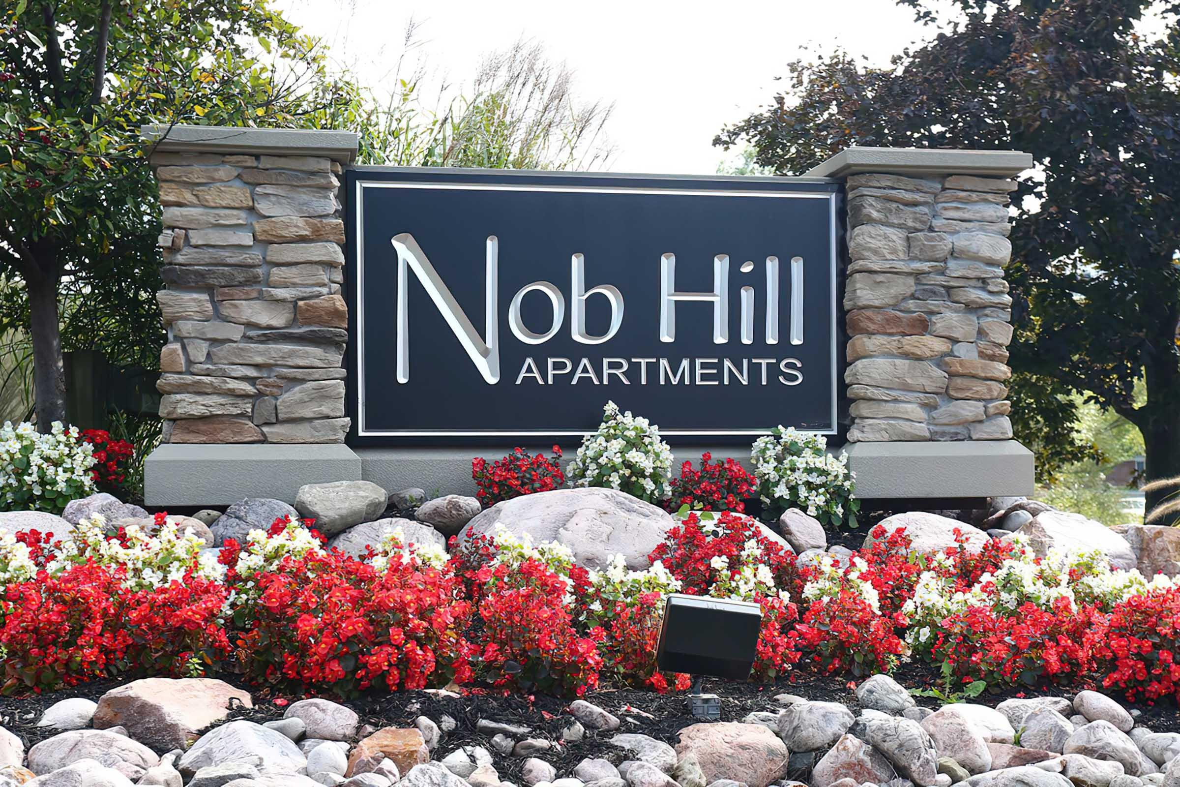 Sign for Nob Hill Apartments, featuring the name prominently displayed in white letters against a black background. The sign is framed with stone pillars and surrounded by vibrant red and white flowers, along with decorative rocks, enhancing the entrance's aesthetic appeal.
