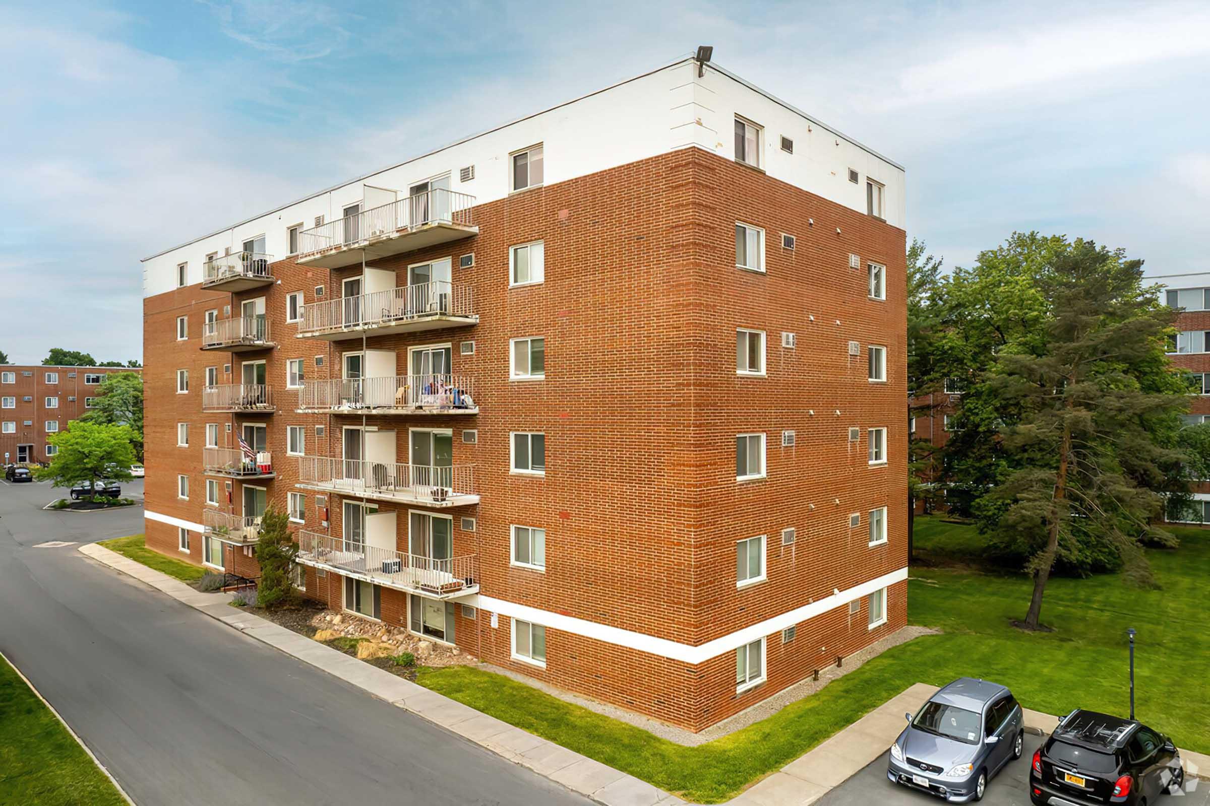A four-story brick apartment building with balconies, surrounded by green lawns and trees. A paved pathway runs along the building, and parked cars are visible in the foreground. The sky is partly cloudy, adding to the scenic view of the residential area.