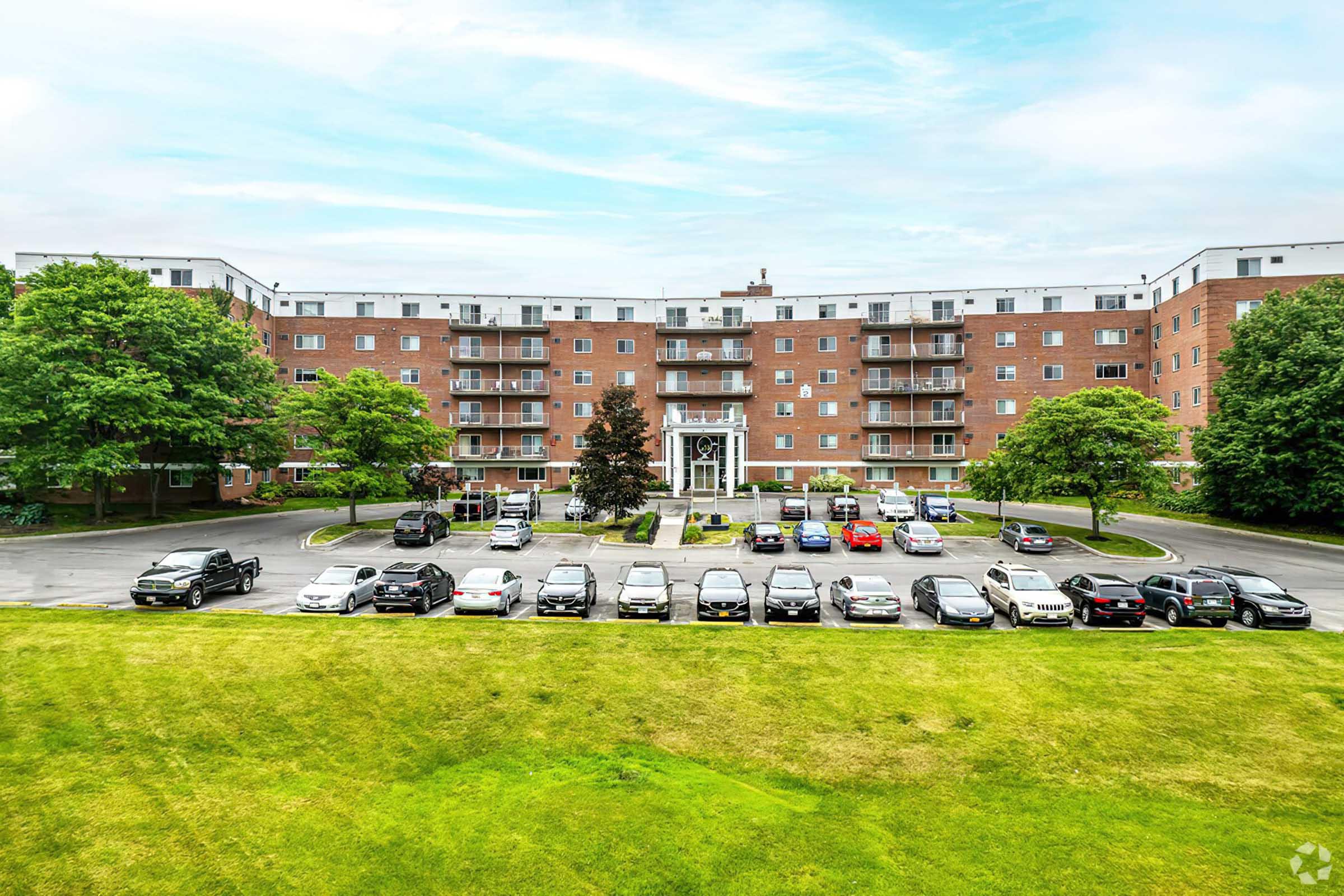 A multi-story brick apartment building surrounded by greenery and a parking lot. Several parked cars are visible in front of the building, with trees and lawn space adding to the landscape. The sky is clear with some clouds, creating a bright atmosphere.