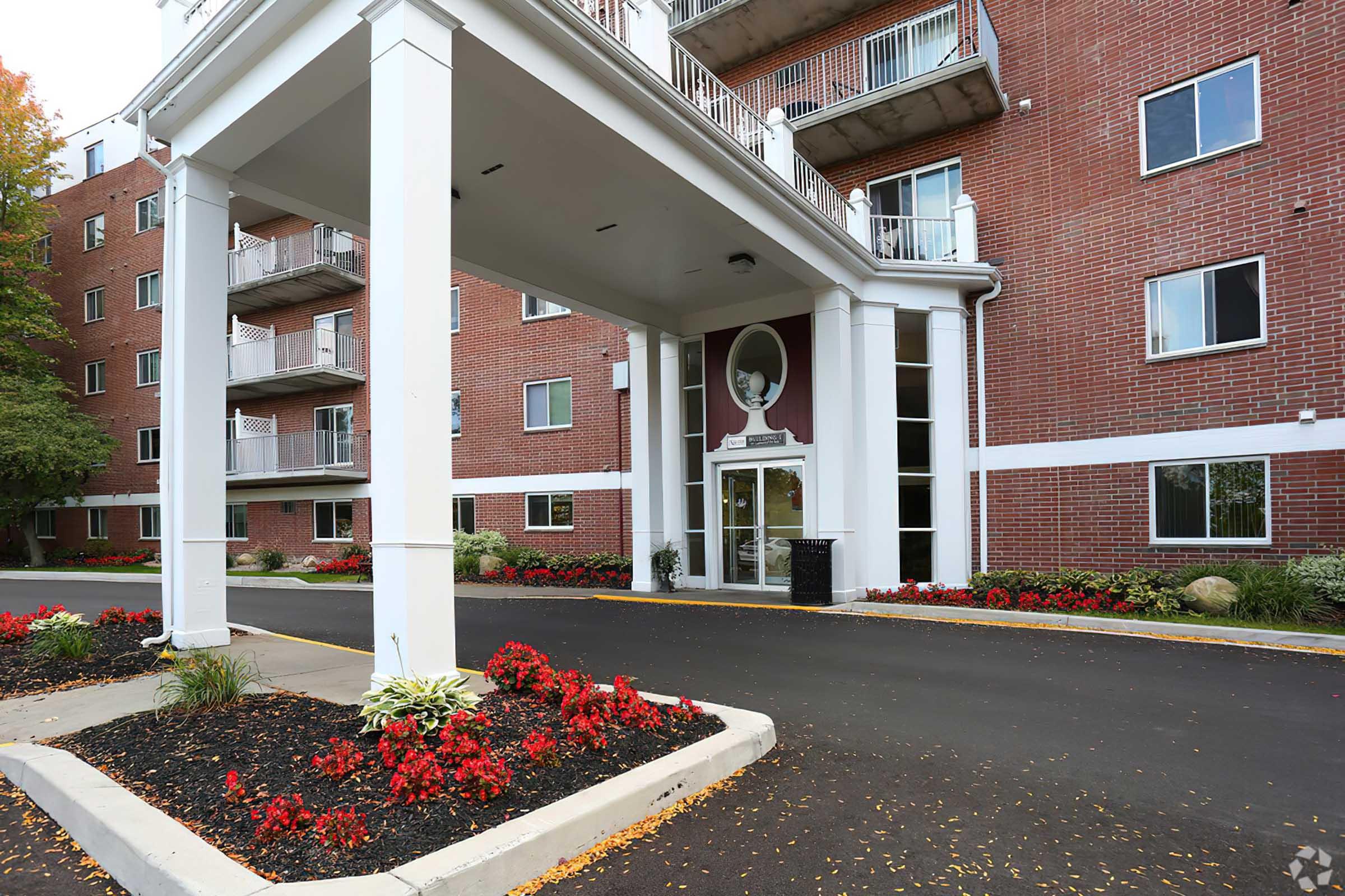 Exterior view of a multi-story brick building entrance, featuring a covered porch supported by white pillars. Colorful flower beds with red flowers line the walkway, and the building has balconies on the upper floors. A parking area is visible in front.
