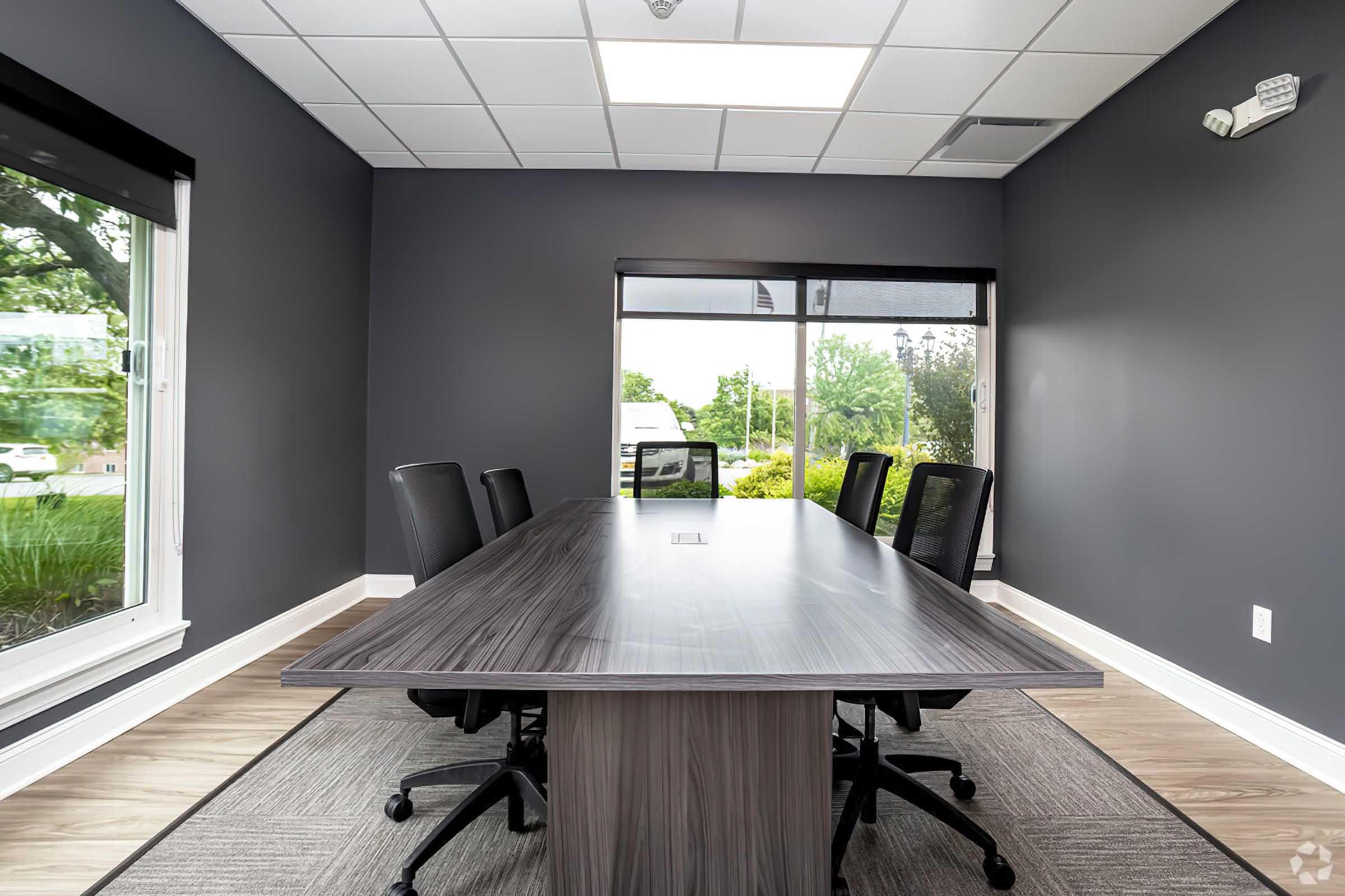 A modern conference room featuring a large wooden table surrounded by black office chairs. Large windows allow natural light to enter, with greenery visible outside. The walls are painted dark gray, and the flooring is a light wood laminate. The room is designed for meetings and collaboration.
