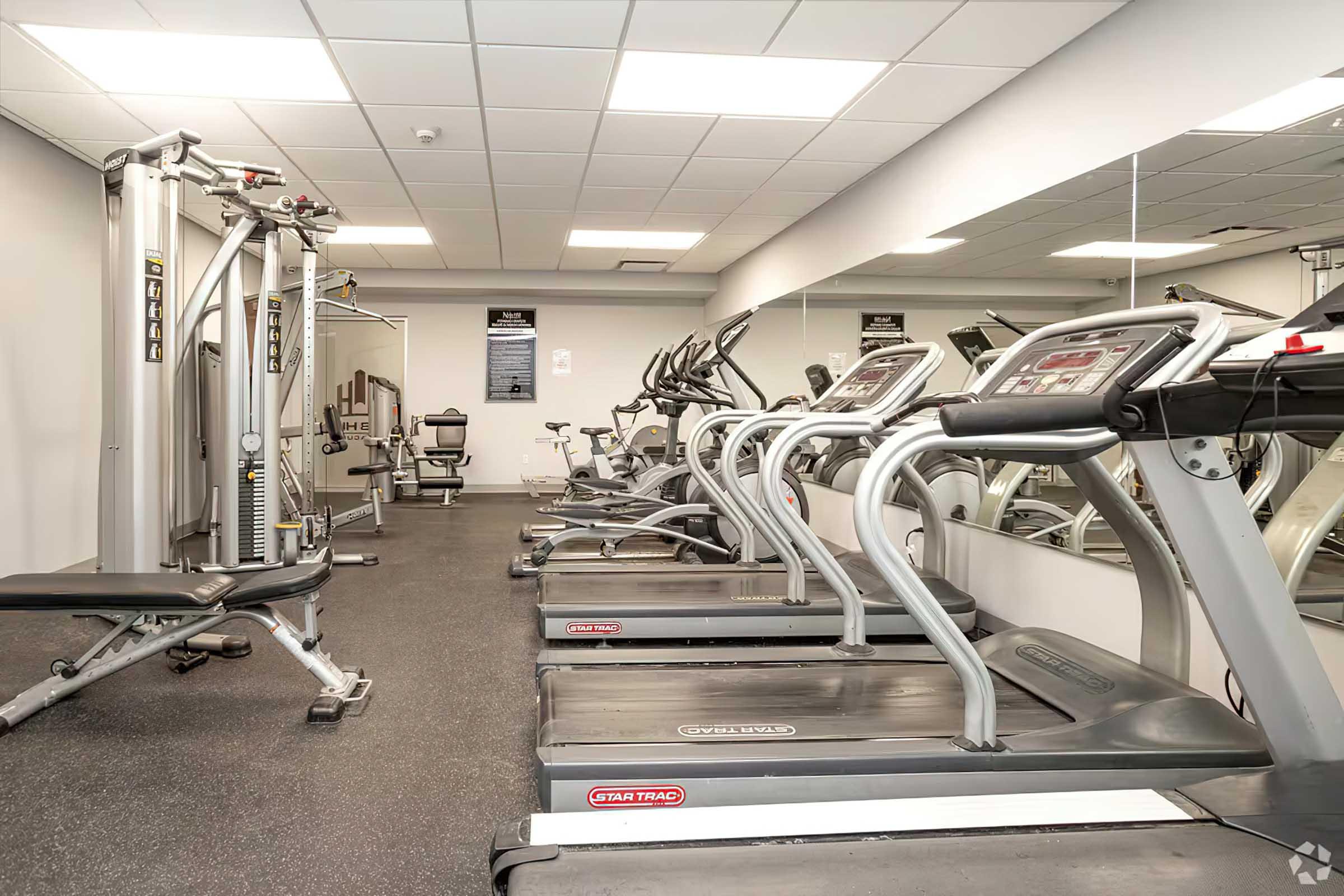 A modern gym interior featuring several exercise machines including treadmills and a multi-gym unit. The space is well-lit with mirrors lining one wall, and fitness equipment is arranged neatly on a black rubber floor, creating a clean and inviting workout environment.