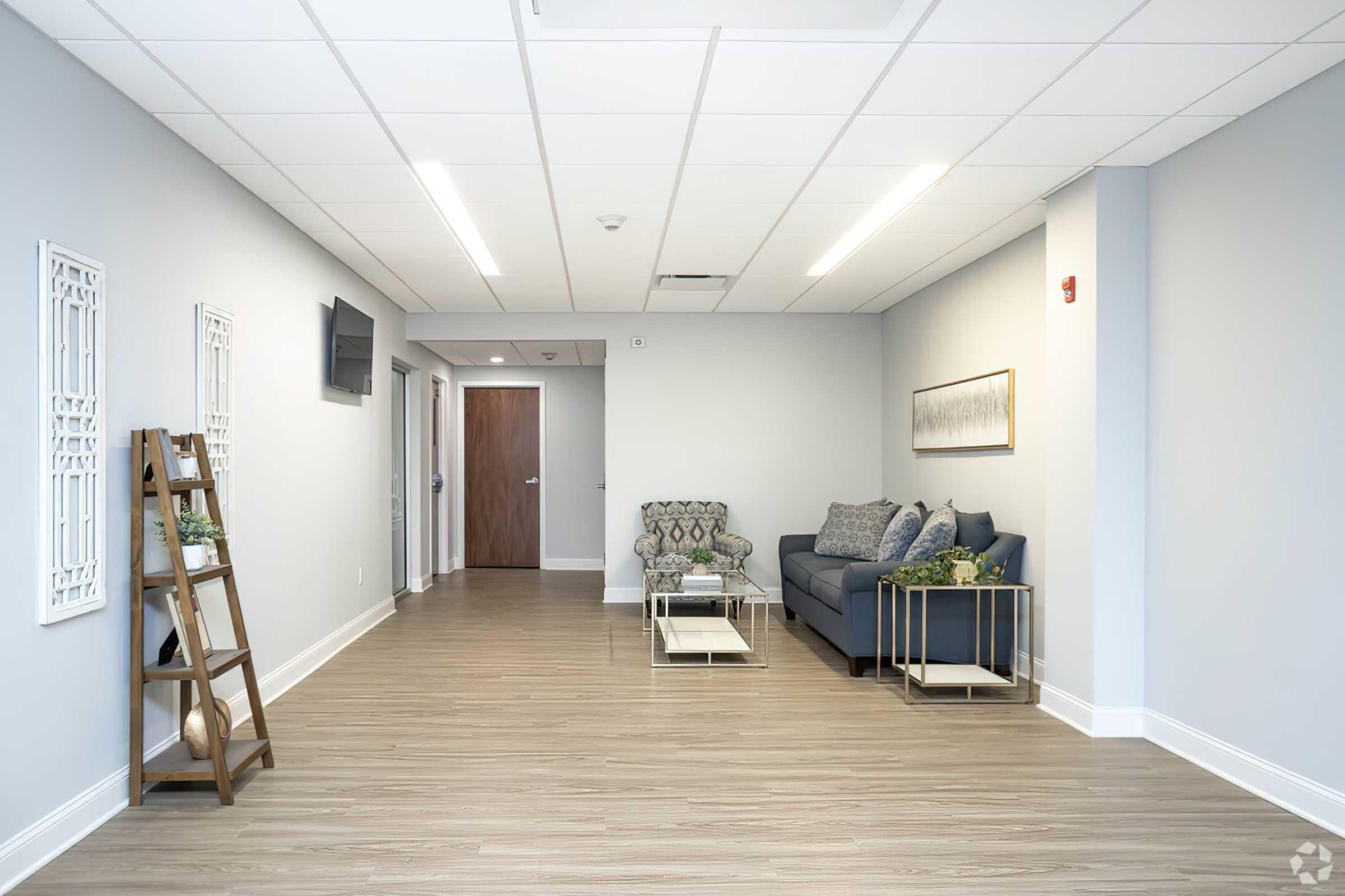 A modern, spacious lounge area with light gray walls and wood flooring. It features a blue sofa with decorative cushions, a ladder shelf with a vase, and a minimalist wall art piece. A television is mounted on the wall, and there is a doorway in the background leading to another room.