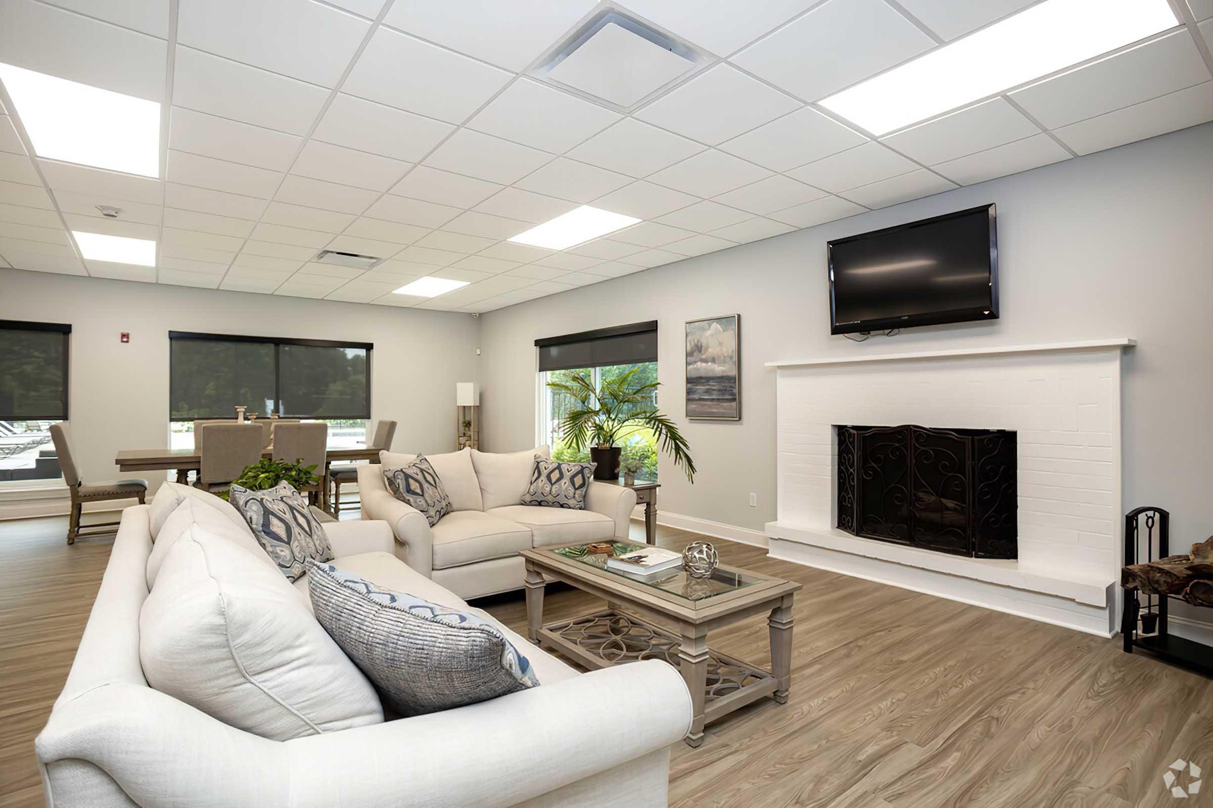 A spacious and modern living room featuring a light-colored couch with decorative pillows, a wooden coffee table, a fireplace with an ornate screen, and a wall-mounted TV. Large windows allow natural light to flow in, with a dining area visible in the background, designed for comfort and relaxation.