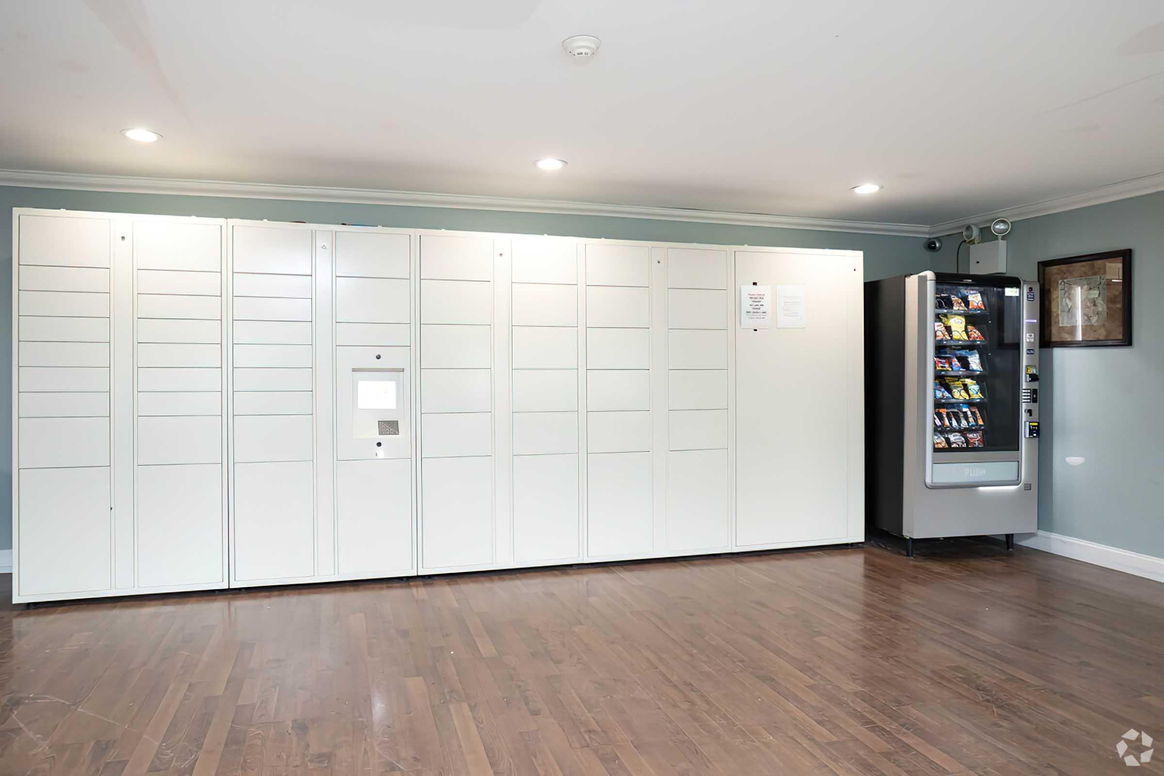 A room featuring a row of white package lockers on the left and a vending machine on the right. The floor is wooden, and the walls are painted light blue with a framed picture hanging on the wall. The space is well lit with overhead lights.
