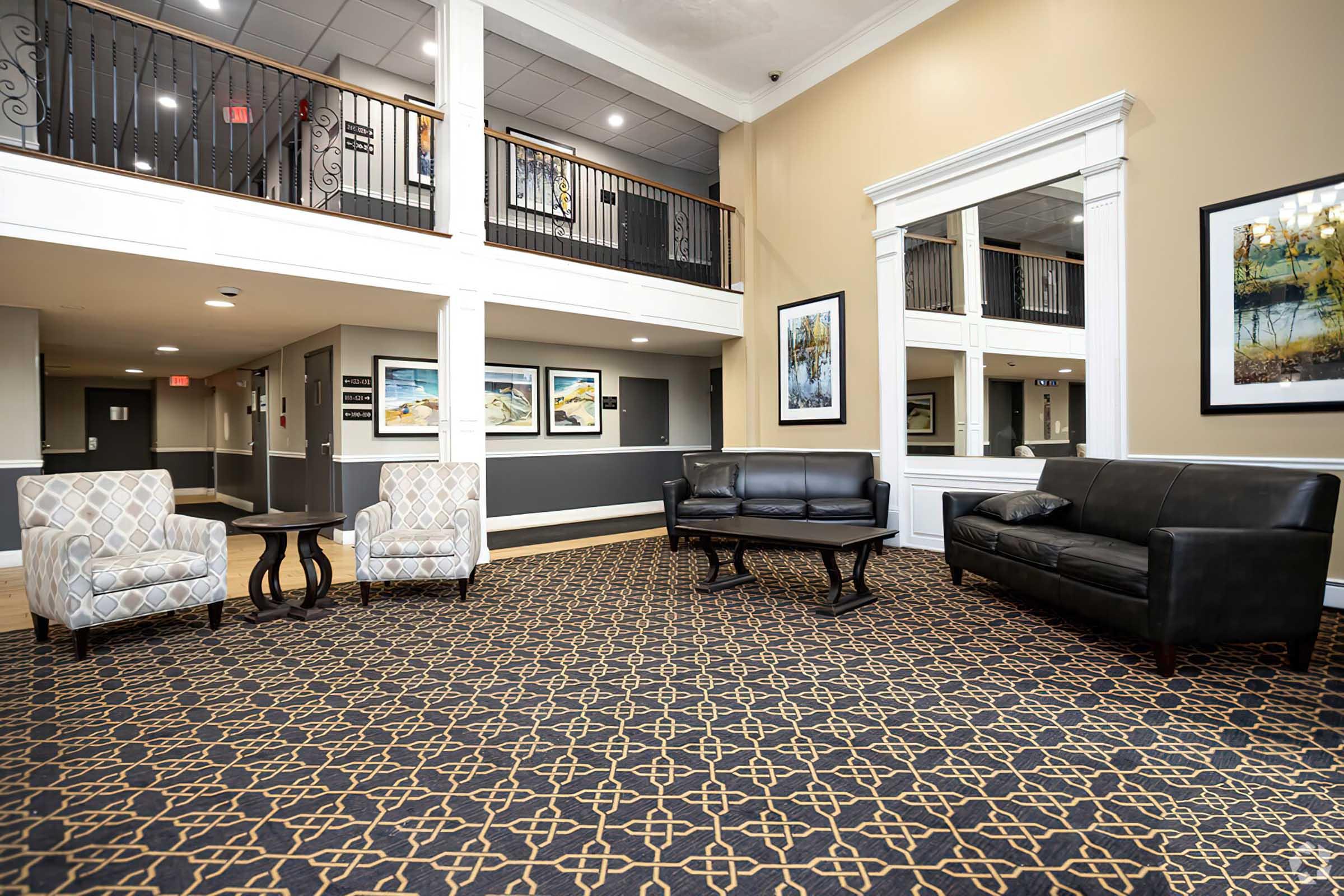 A modern interior space featuring a patterned carpet, two black leather sofas, and a small table. The area has elegant decor with framed artwork on the walls, large windows providing natural light, and a welcoming atmosphere ideal for relaxation or socializing.