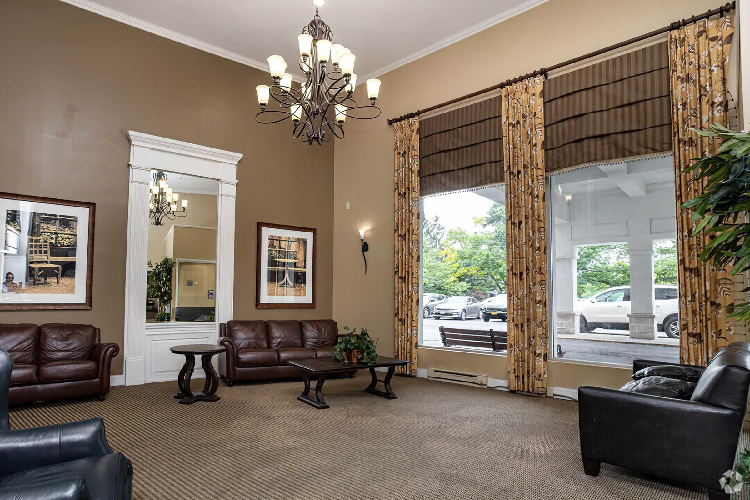 Cozy lobby area featuring two leather sofas, a coffee table, and a large chandelier. The walls are painted a warm brown, adorned with framed artwork. Large windows with decorative curtains allow natural light to fill the space, showcasing greenery outside. A mirror reflects the room’s inviting atmosphere.