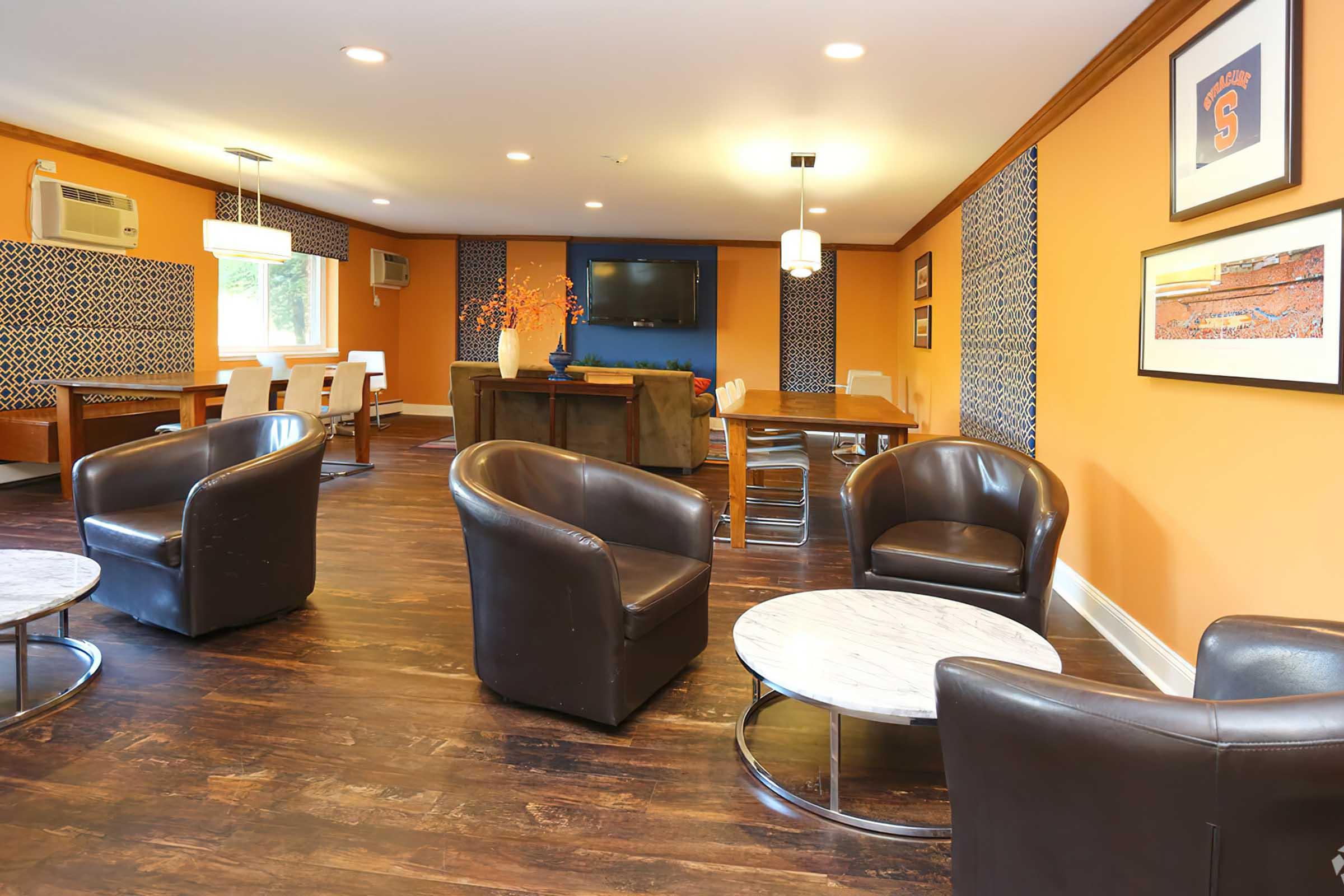 A cozy interior space featuring modern furniture. Two round coffee tables are surrounded by comfortable leather armchairs. In the background, there is a dining area with white chairs and a large television mounted on the wall. The walls are painted orange with patterned accent sections, and the flooring is dark wood.