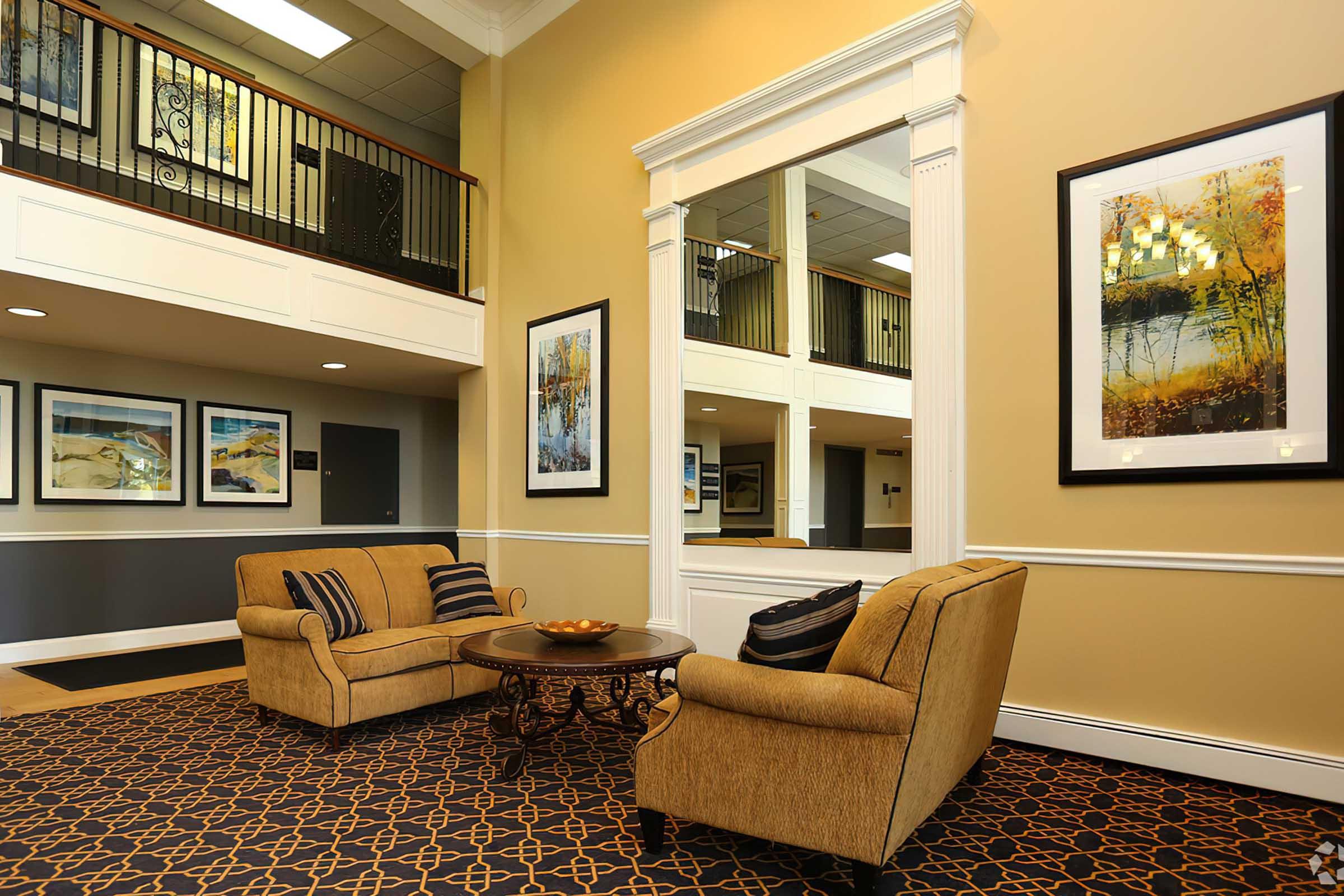 A cozy lobby area featuring two upholstered chairs with striped cushions and a round coffee table, surrounded by decorative artwork on the walls. The space is brightly lit with large mirrors and elegant interior decor, creating an inviting atmosphere.