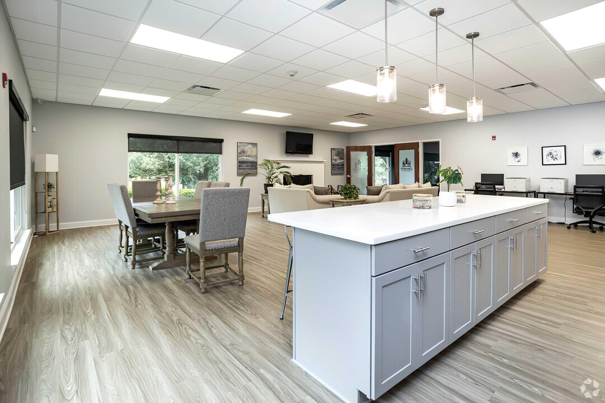 A modern communal space featuring a dining table with gray chairs, a kitchen island with storage, and comfortable seating areas. Large windows allow natural light, and the decor includes neutral tones and contemporary furnishings. The environment is inviting and spacious, suitable for gatherings or meetings.