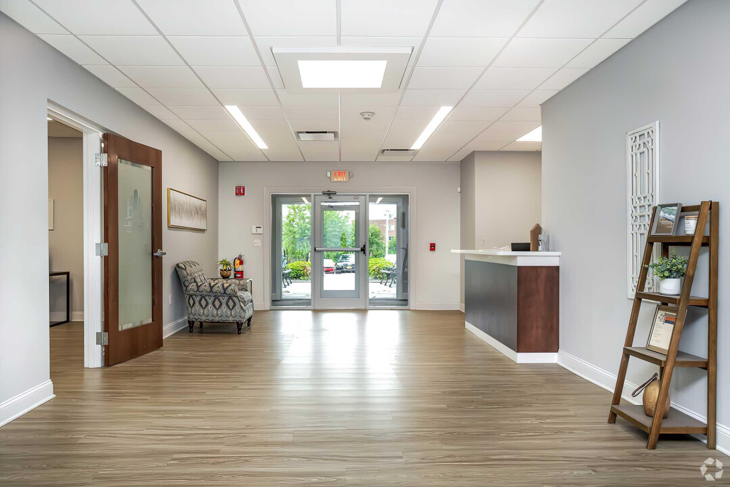 A modern reception area featuring a light gray color scheme, hardwood floors, and a welcoming atmosphere. There is a comfortable chair to the left, a wooden reception desk ahead, and double glass doors leading outside. A decorative ladder shelf on the right holds potted plants and decorative items.