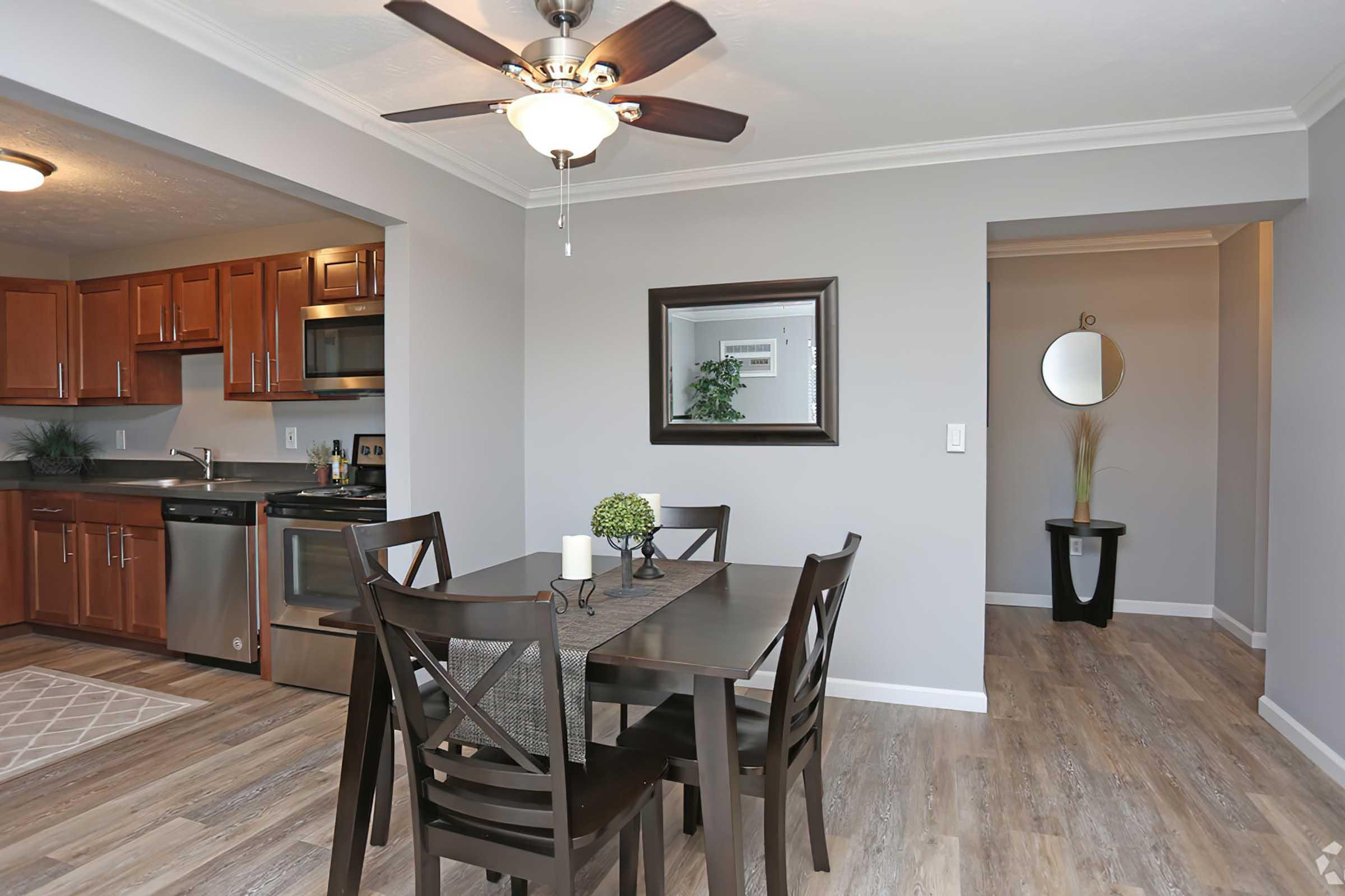 A modern dining area with a wooden table and chairs, featuring a candle centerpiece. To the left, a sleek kitchen with wooden cabinets and stainless steel appliances. A neutral-colored wall is adorned with a large mirror, while a small entryway with a round mirror and decorative plant is visible on the right.