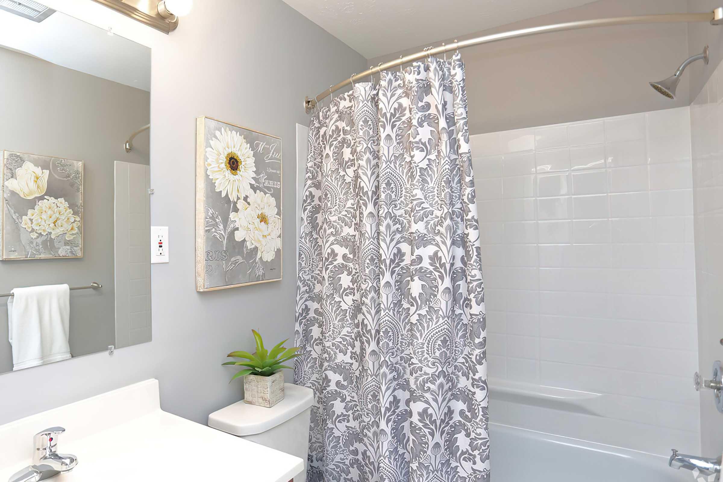 A modern bathroom featuring a white bathtub and shower with a decorative gray and white shower curtain. There's a small potted plant on the bathroom counter, a wall art piece with white flowers, and a towel hanging on the wall. The walls are painted in a light gray color, creating a clean and elegant atmosphere.