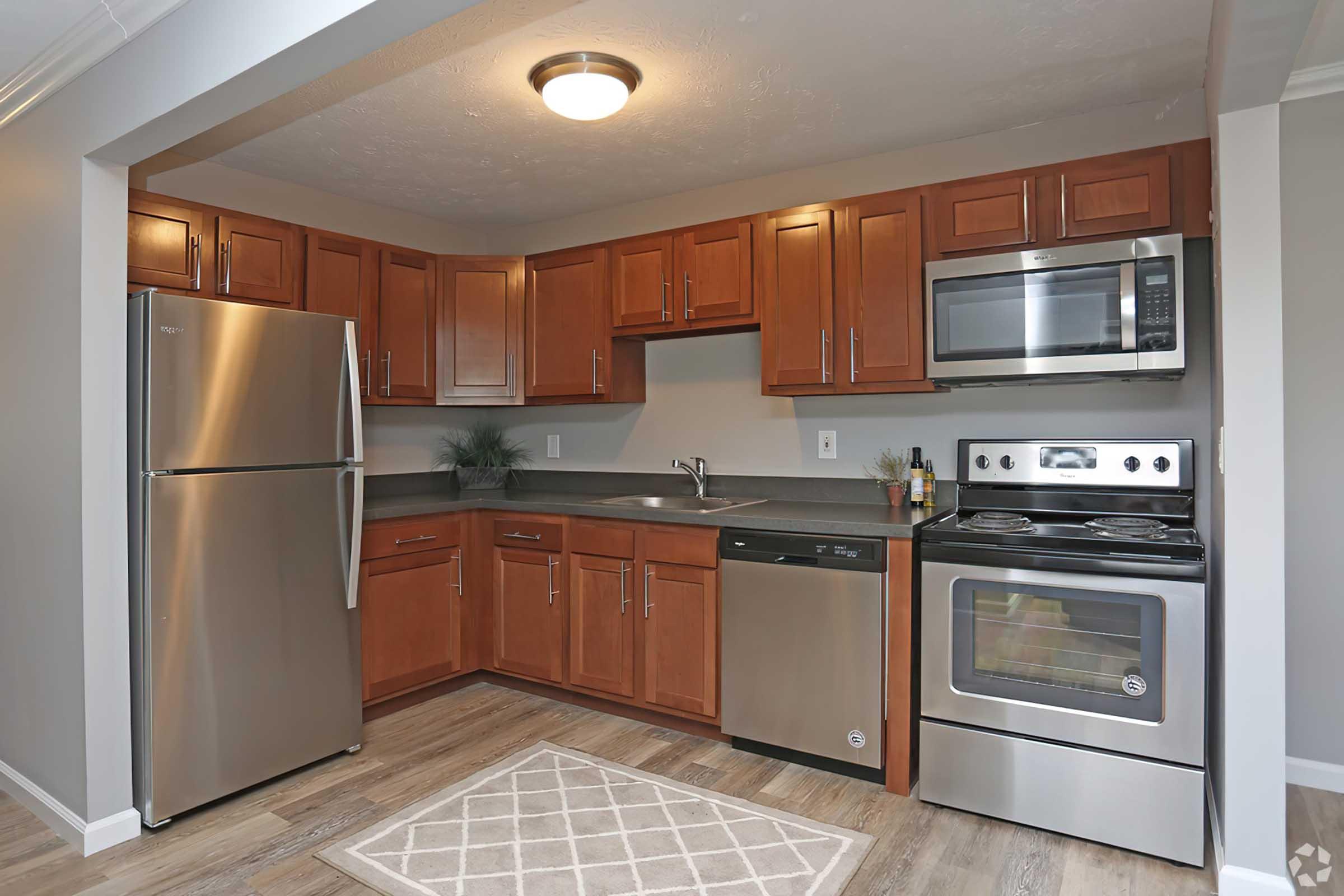 A modern kitchen featuring stainless steel appliances, including a refrigerator, microwave, and stove. The cabinetry is wooden with a rich brown finish, complemented by a gray countertop. A small area rug is placed on the floor, and there is a potted plant on the countertop for decoration.