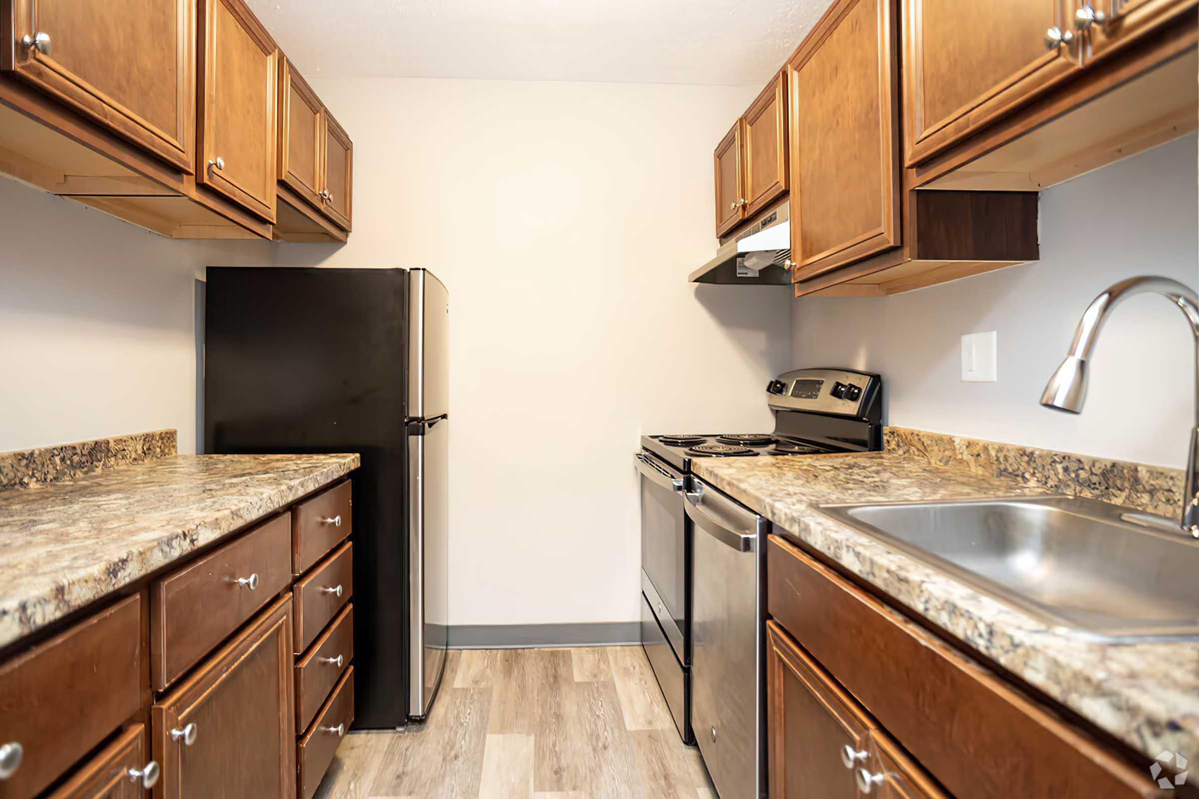 Modern kitchen featuring wooden cabinets, a black refrigerator, stainless steel stove and oven, and a stainless steel sink. Countertops are made of speckled granite, and the flooring is light-colored laminate. The wall is painted a neutral tone, contributing to a clean and inviting atmosphere.