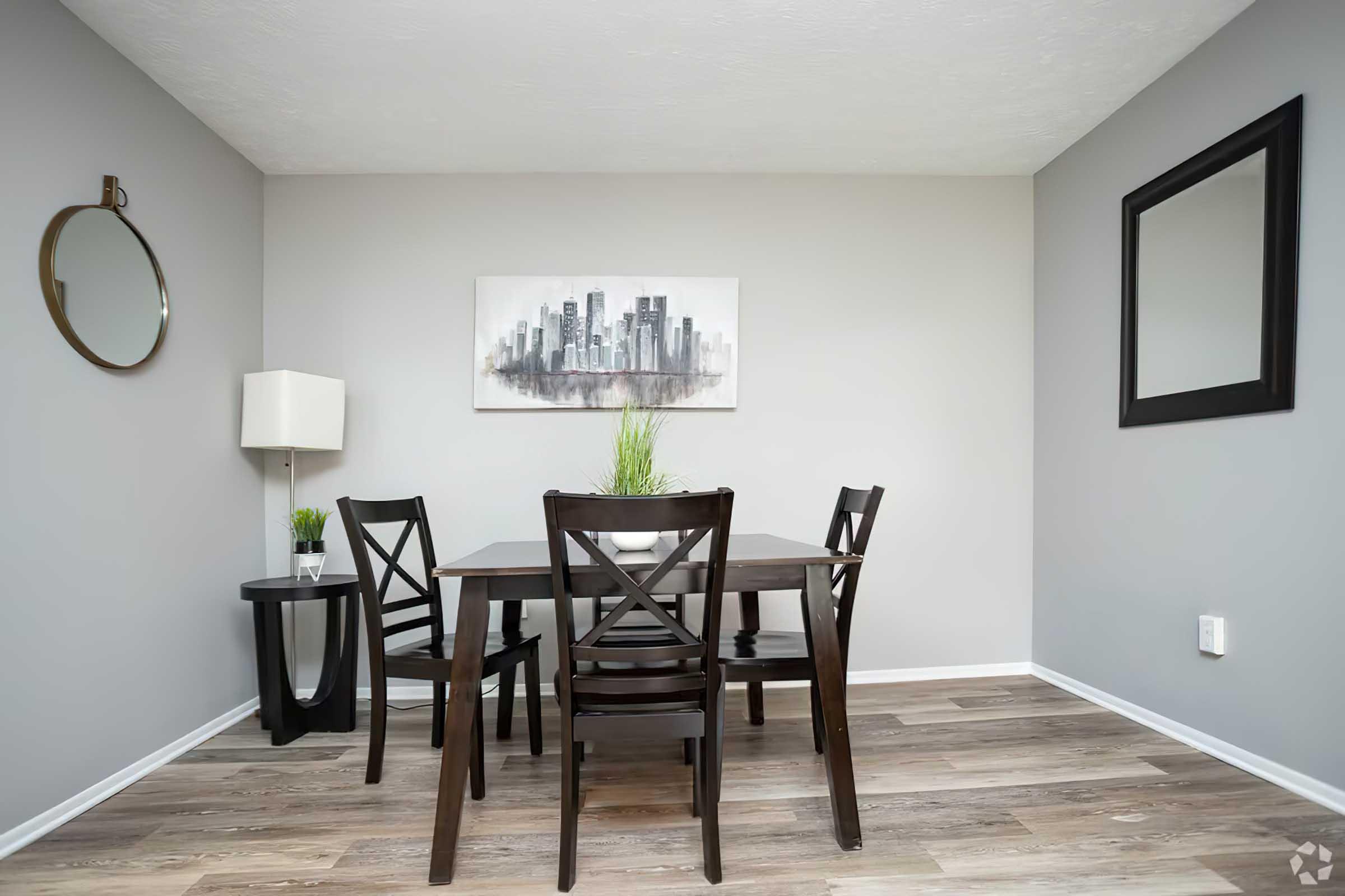 A modern dining area featuring a dark wooden table with four chairs, a round mirror on the left wall, and a lamp beside a small black side table. The walls are painted light gray, and a cityscape painting hangs above the table, complemented by a small potted plant. The flooring is a warm wood finish.