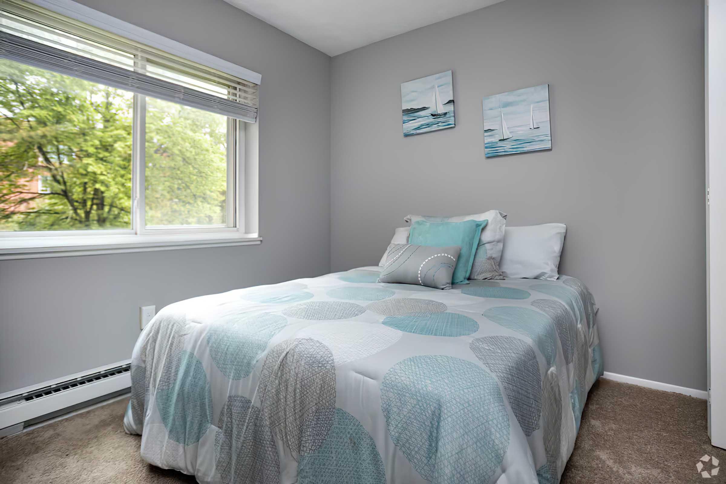 A cozy bedroom featuring a double bed with a circular patterned blanket, two decorative pillows, and light gray walls. There are three framed pictures of boats on the wall, and a window allows natural light to fill the room, overlooking greenery outside. The carpet is a neutral color.