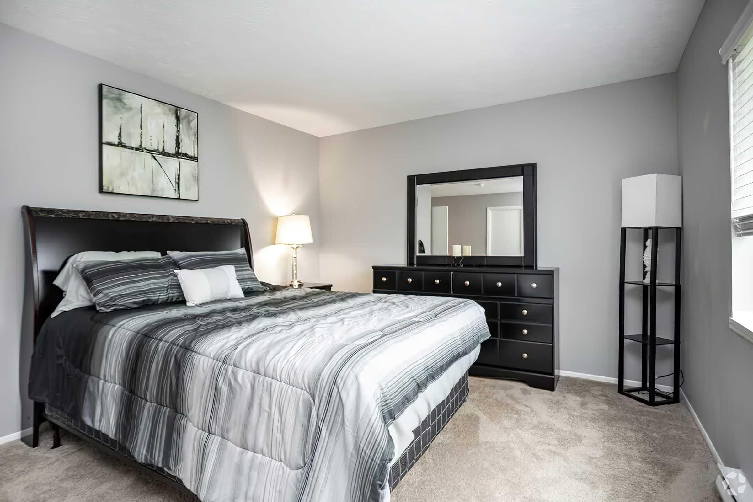 A well-lit bedroom featuring a king-sized bed with gray and white striped bedding, a modern black dresser with gold knobs, a sleek black mirror, and a bedside lamp. The walls are painted gray, and there is a tall, narrow bookshelf with a white lamp beside the window.