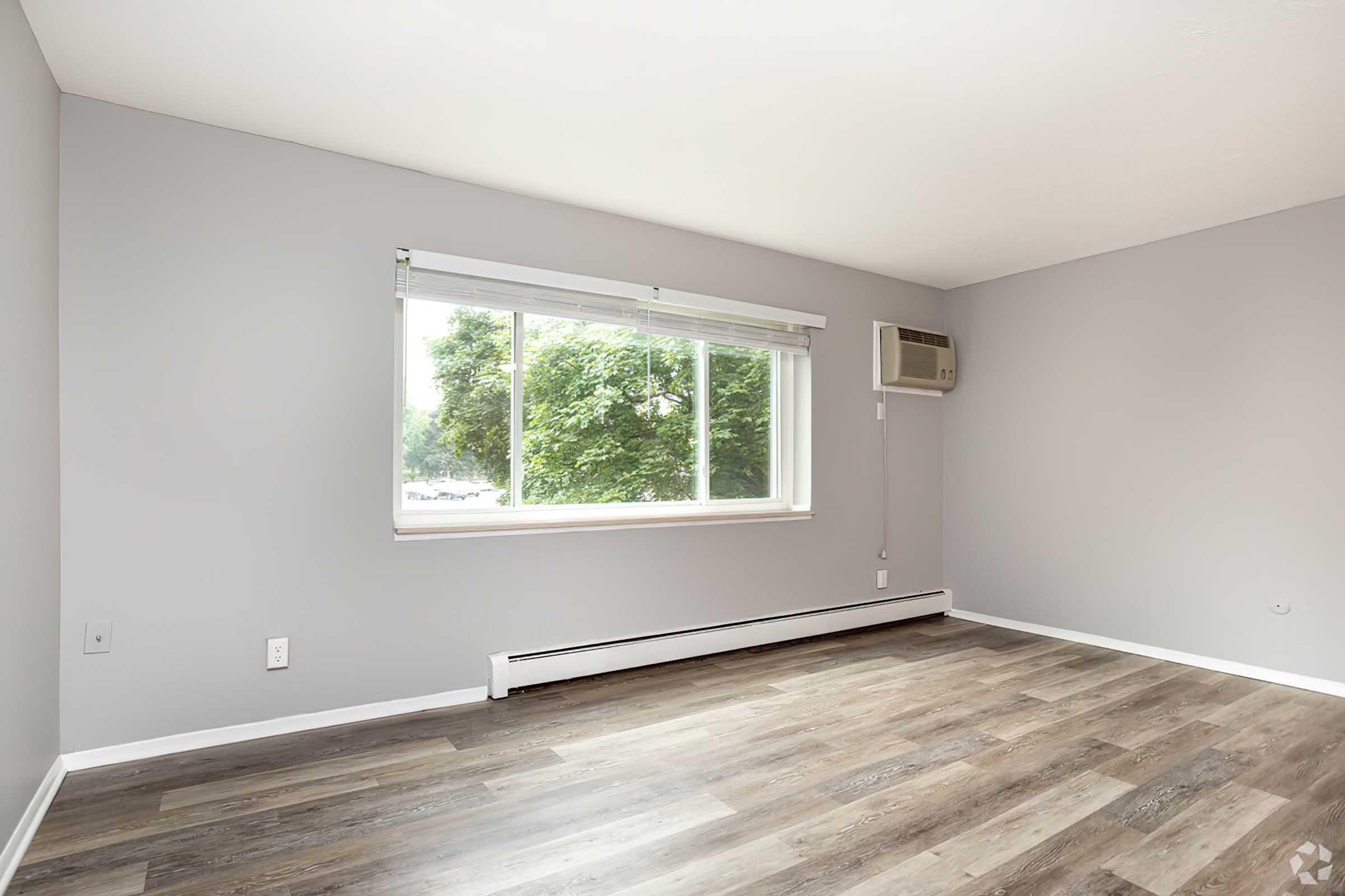A bright, empty room with gray walls and laminate flooring. A large window with white blinds allows natural light to enter, and an air conditioning unit is mounted on the wall. The space appears clean and modern, ready for furnishings.