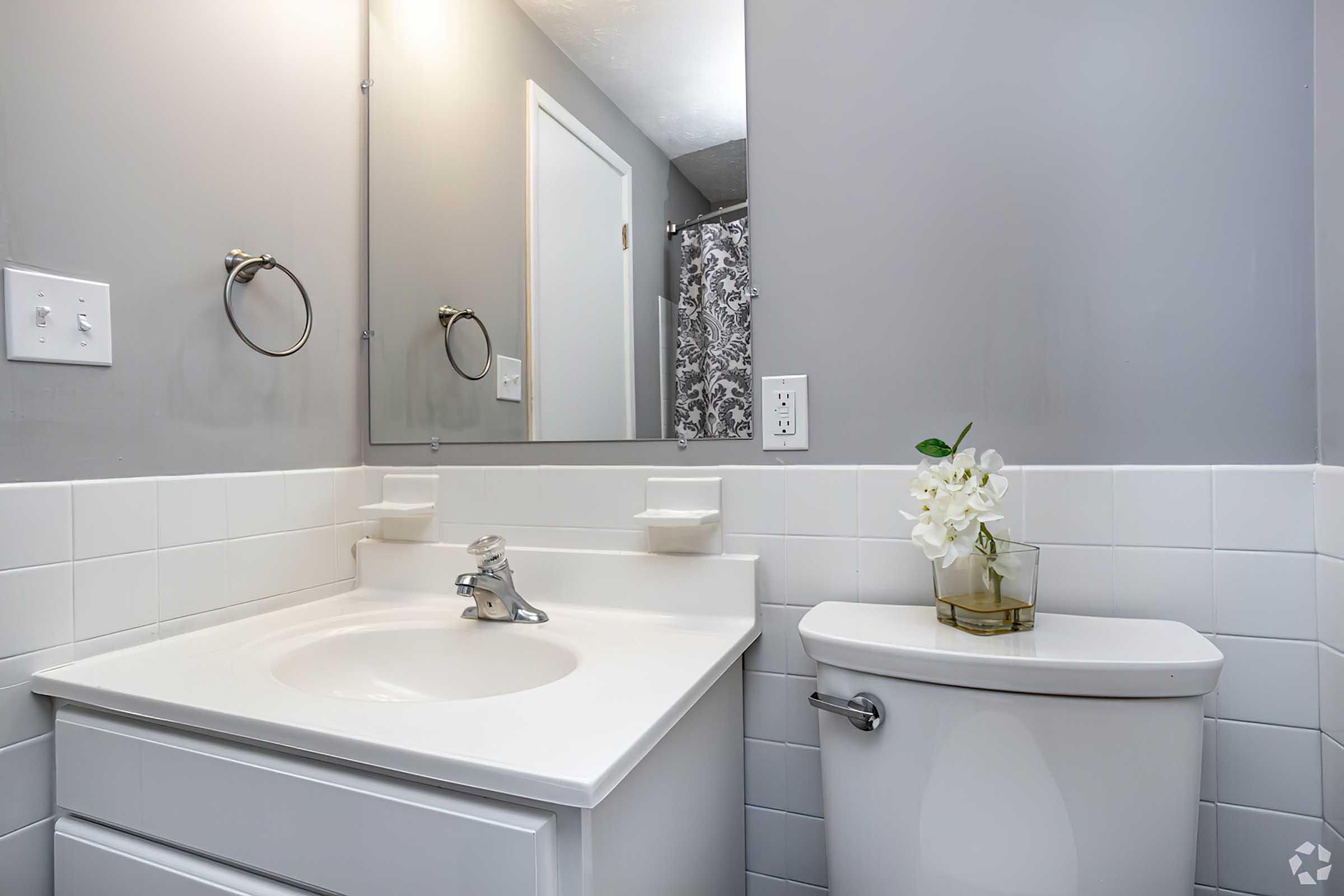 A clean and modern bathroom featuring a white sink and countertop, a matching toilet, a wall-mounted mirror, and a towel holder. The walls are painted in a soft gray color, with white tiled accents. A small vase with a white flower adds a touch of decor to the countertop.