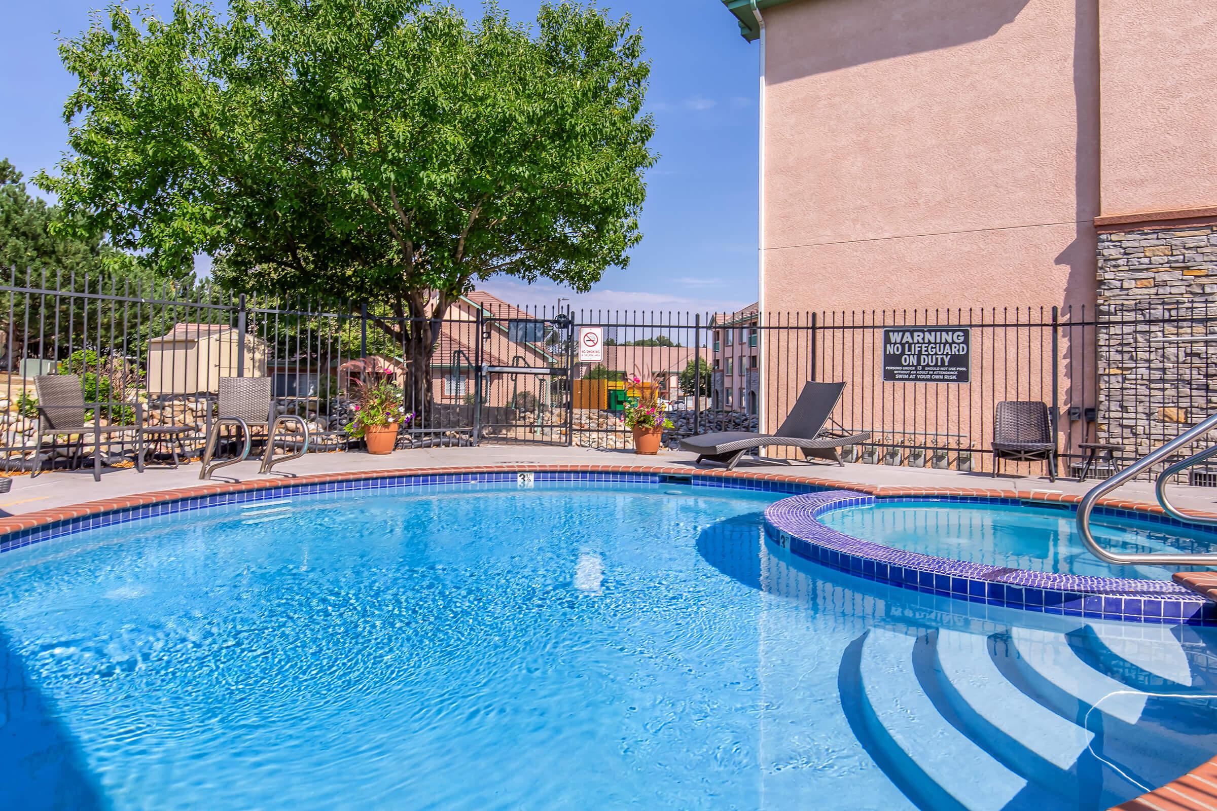 A clear blue swimming pool with a curved jacuzzi section, surrounded by lounge chairs and potted plants. A large tree provides shade nearby, and a sign indicating pool rules is visible on the fence. The scene is bright and inviting, reflecting a sunny day.