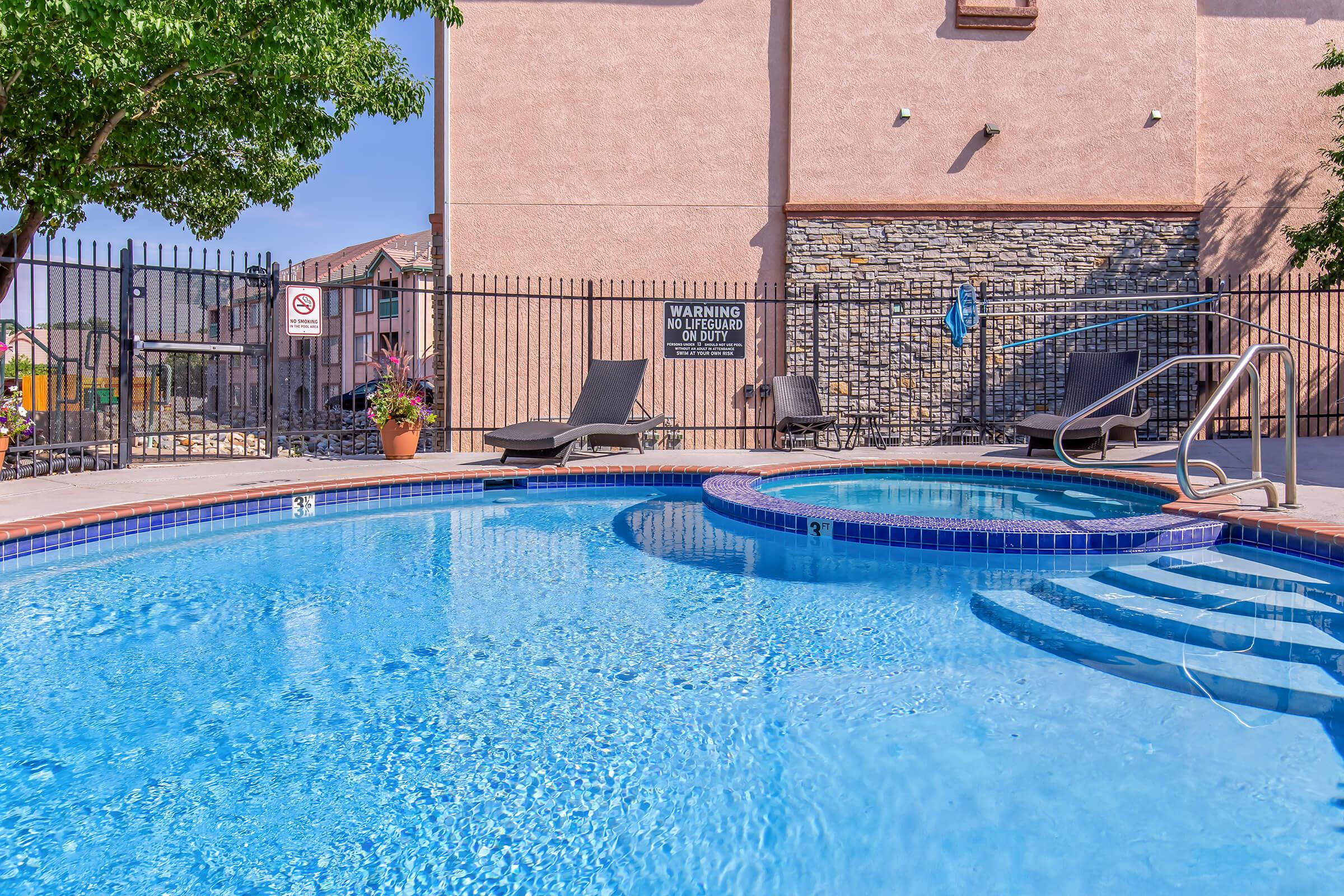 A clear blue swimming pool with a spa area, surrounded by a black metal fence. There are two lounge chairs placed by the pool, and a potted plant nearby. The sun is shining brightly, reflecting off the water's surface, creating a relaxing outdoor atmosphere.