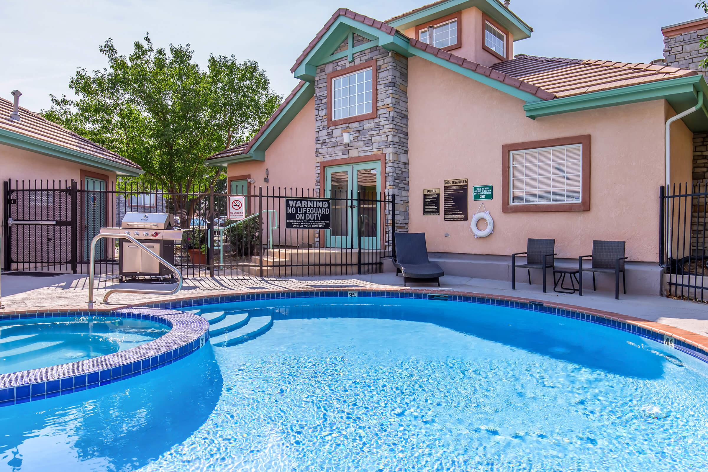 Swimming pool with a hot tub in the foreground, surrounded by a fence. There is a two-story building with a light-colored exterior and stone accents in the background. A warning sign is visible, along with lounge chairs placed nearby. Lush greenery is present in the area, under a clear blue sky.