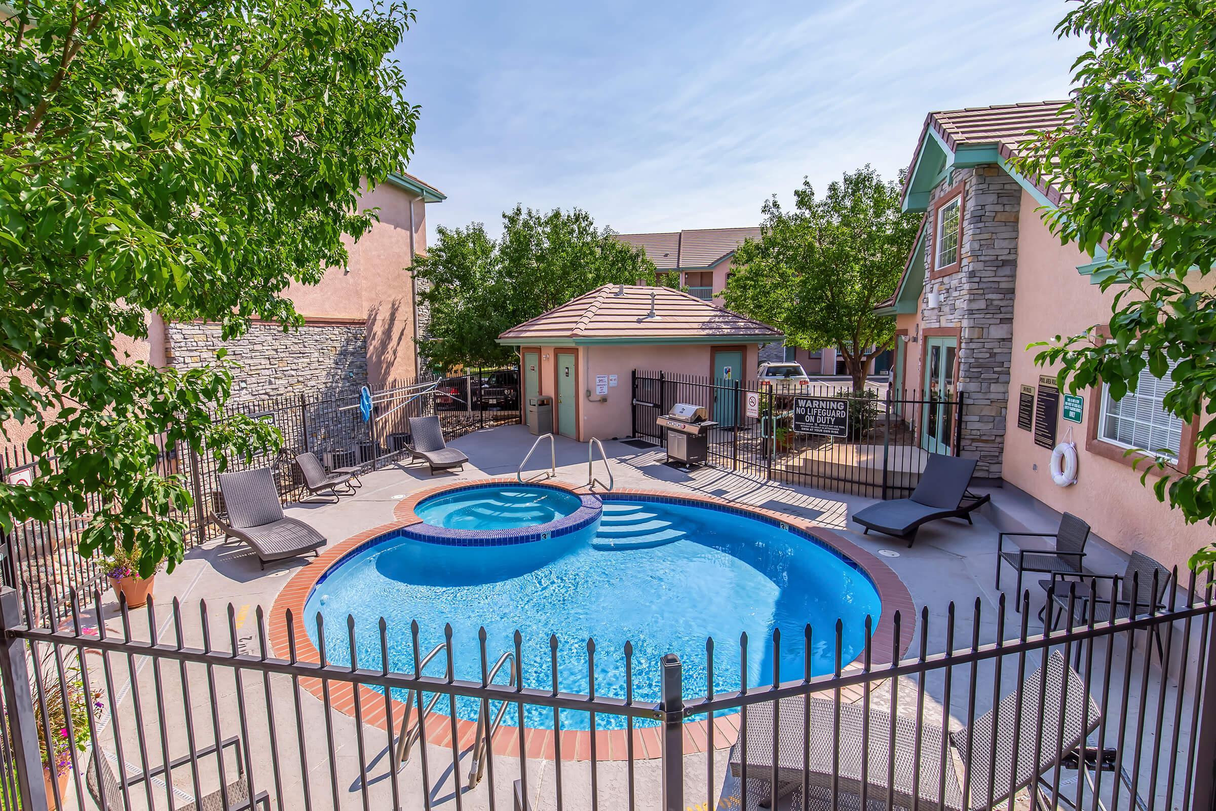 A well-maintained pool area surrounded by a wrought iron fence, featuring a hot tub and several lounge chairs. A small outdoor grill is nearby, and the setting is enhanced by green trees and a clear blue sky. The pool area is part of a residential community.