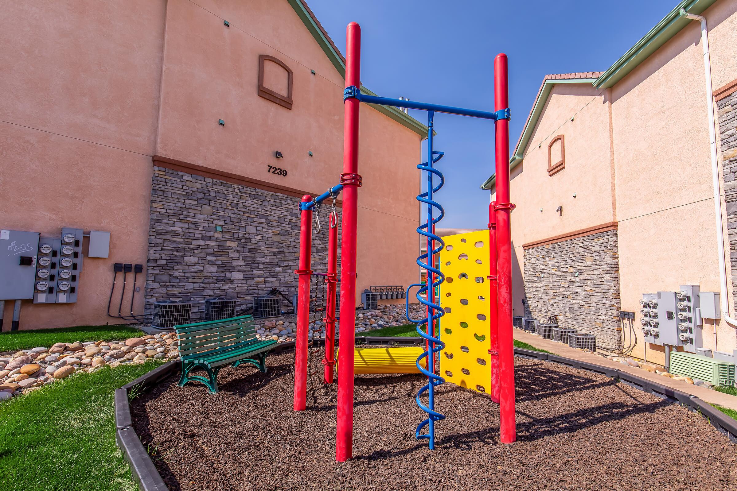 Colorful playground equipment featuring a spiral climbing structure and a large yellow slide surrounded by mulch. Green bench nearby and residential buildings with stone and stucco exteriors in the background. Clear blue sky overhead.