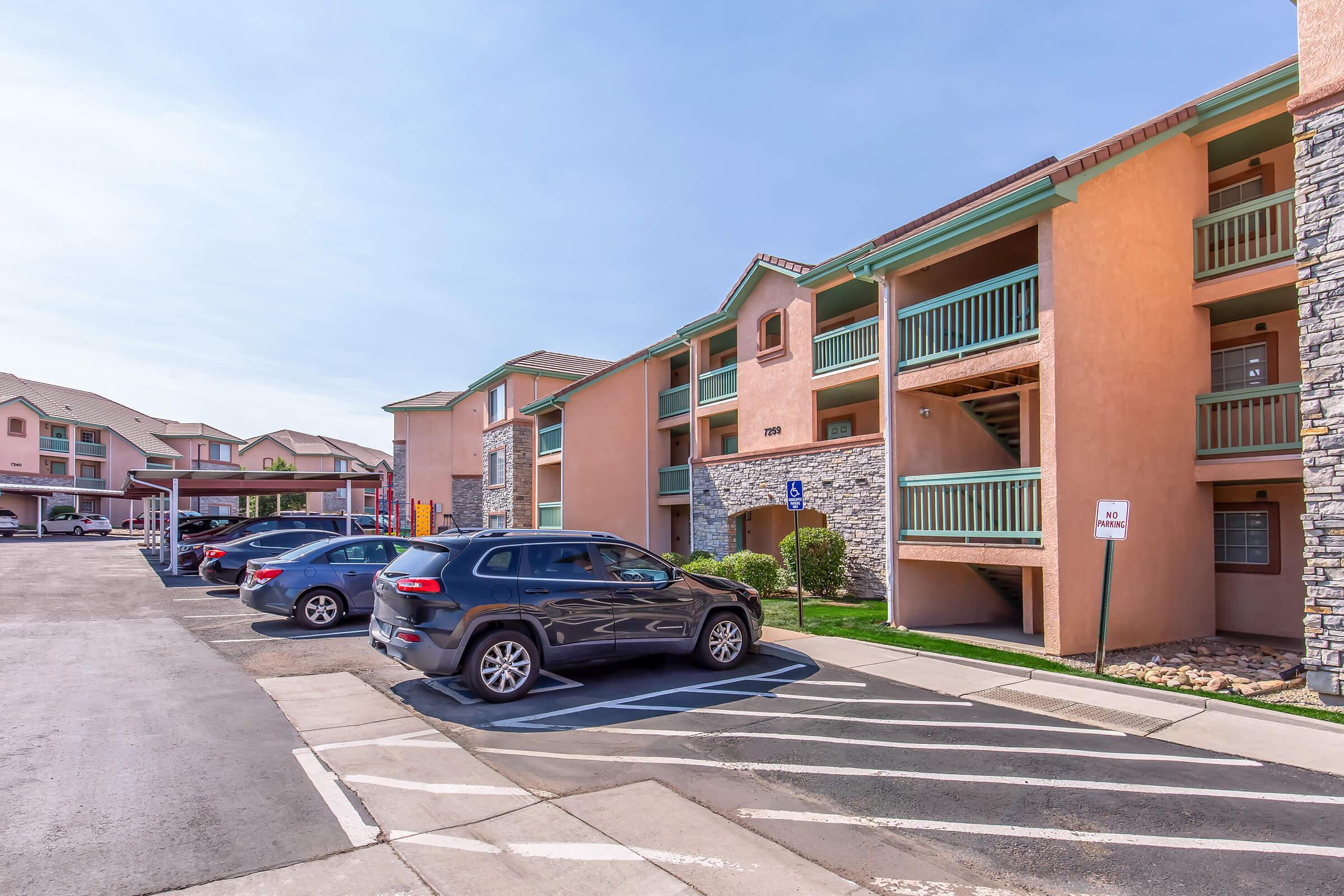 Exterior view of a multi-story apartment complex featuring peach-colored walls, stone accents, and balconies. Several parked cars are visible in front of the building, along with landscaped areas. The sky is clear, indicating a sunny day.