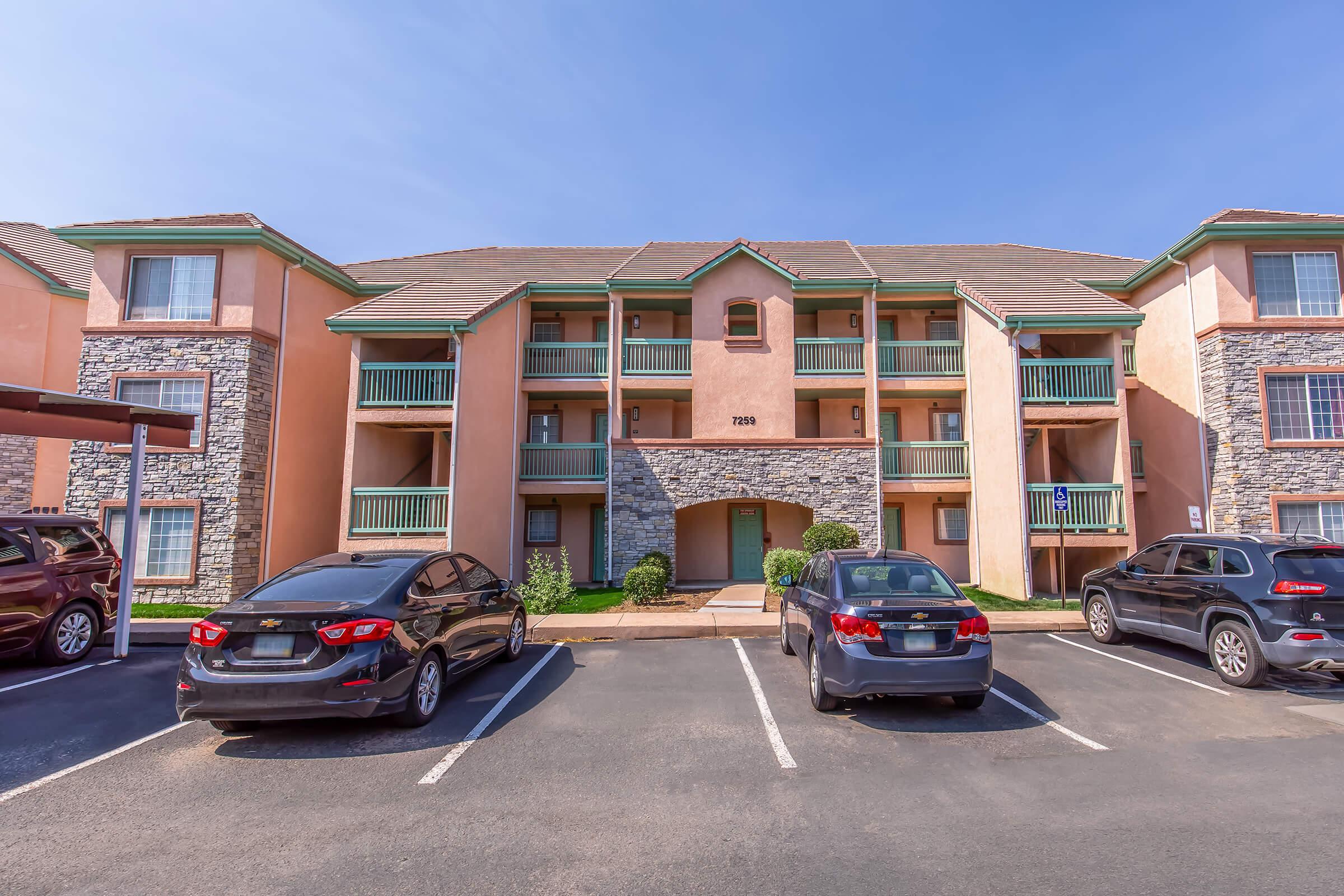 A three-story apartment building featuring a stone facade and pastel-colored walls. Balconies are visible on the second and third floors. Several parked cars are in the foreground, and a clear blue sky is overhead. The setting appears to be a residential area.