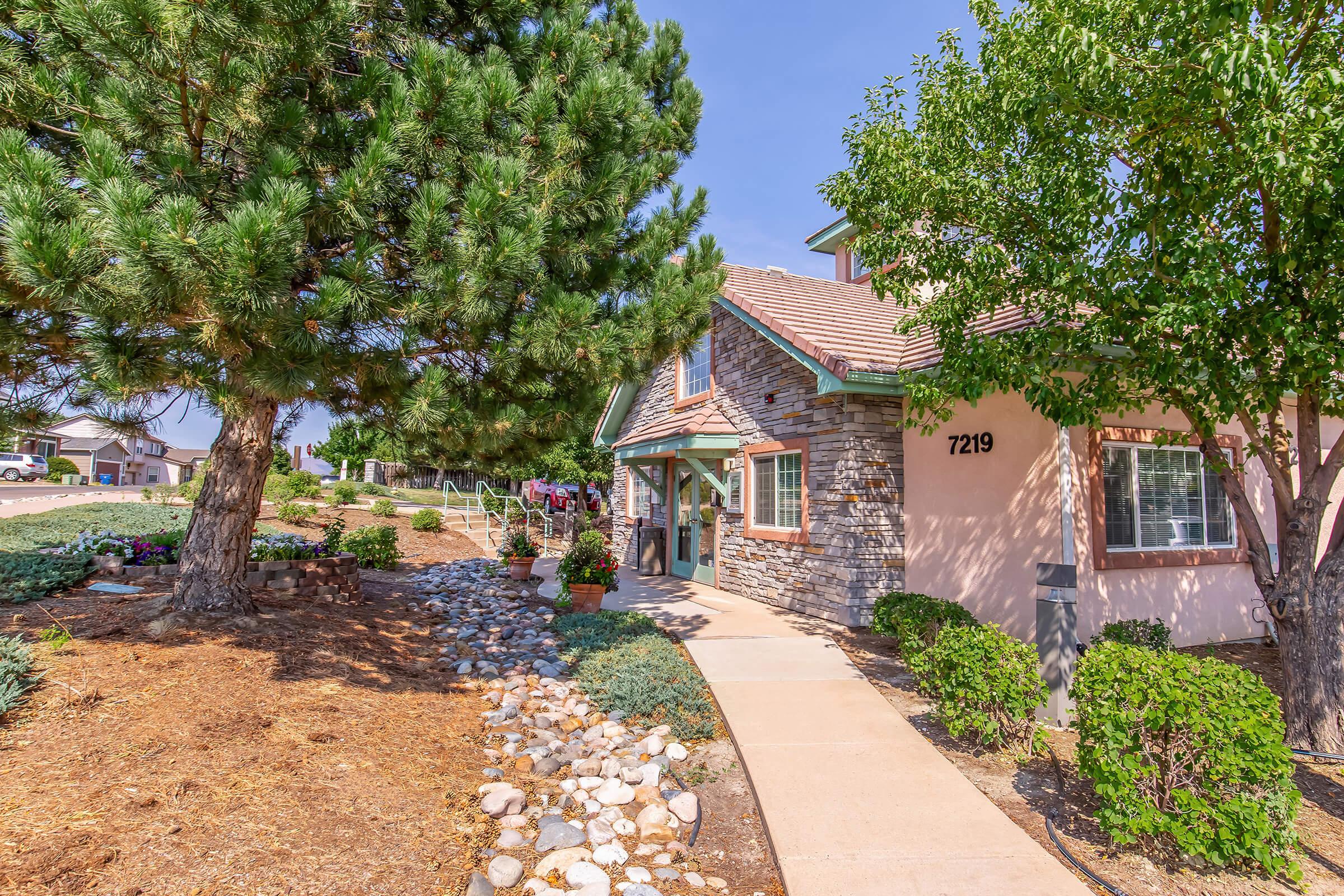 A charming residential entrance featuring a stone-accented house with a sloped roof, surrounded by well-maintained landscaping. A pathway leads to the front door, flanked by greenery and trees. The house number is 7219, and the scene is set against a clear blue sky.