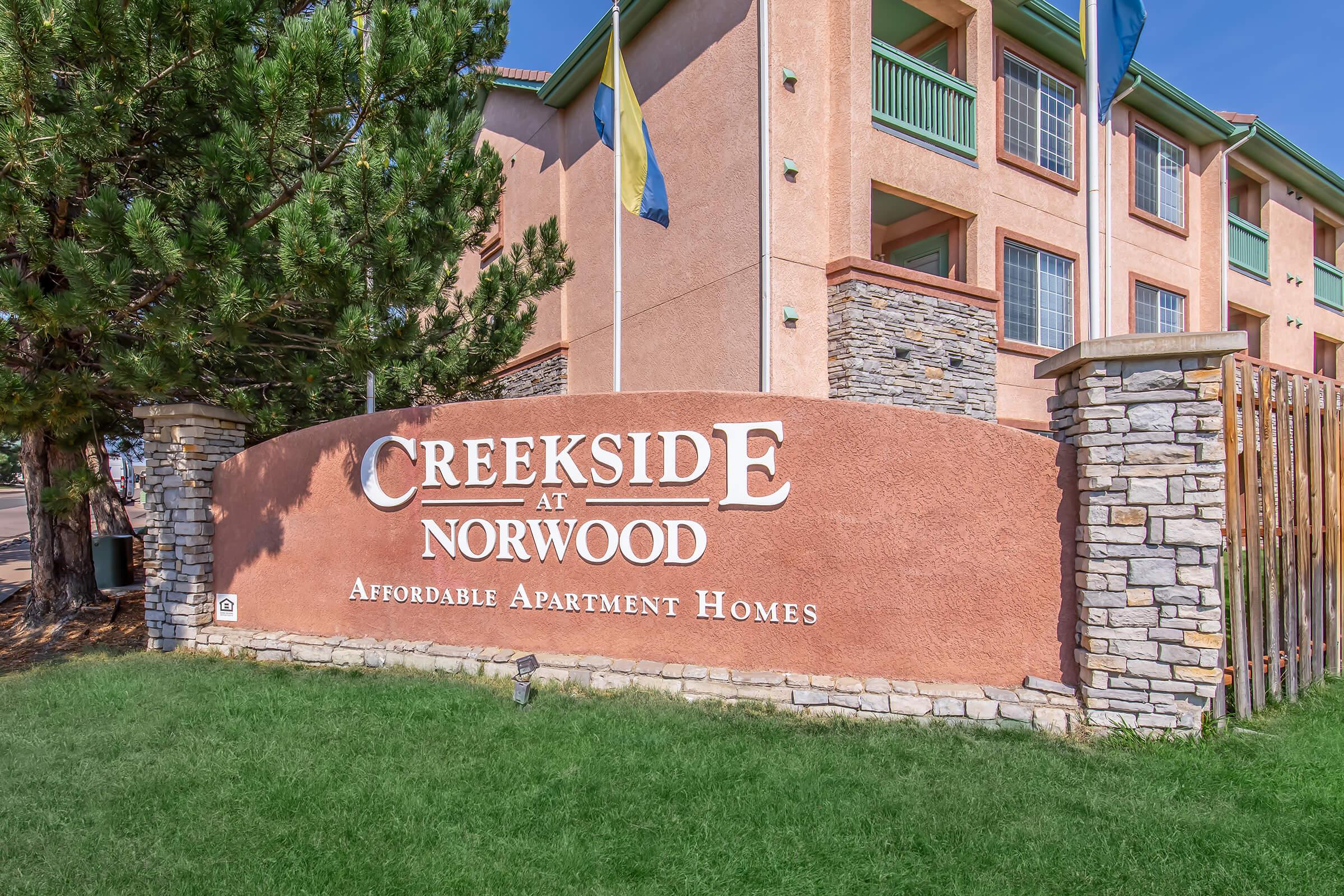 Sign for "Creekside at Norwood," featuring the phrase "Affordable Apartment Homes," set against a backdrop of a multistory building and greenery. Flags are visible, and the design includes stone and stucco elements. The setting suggests a residential area.