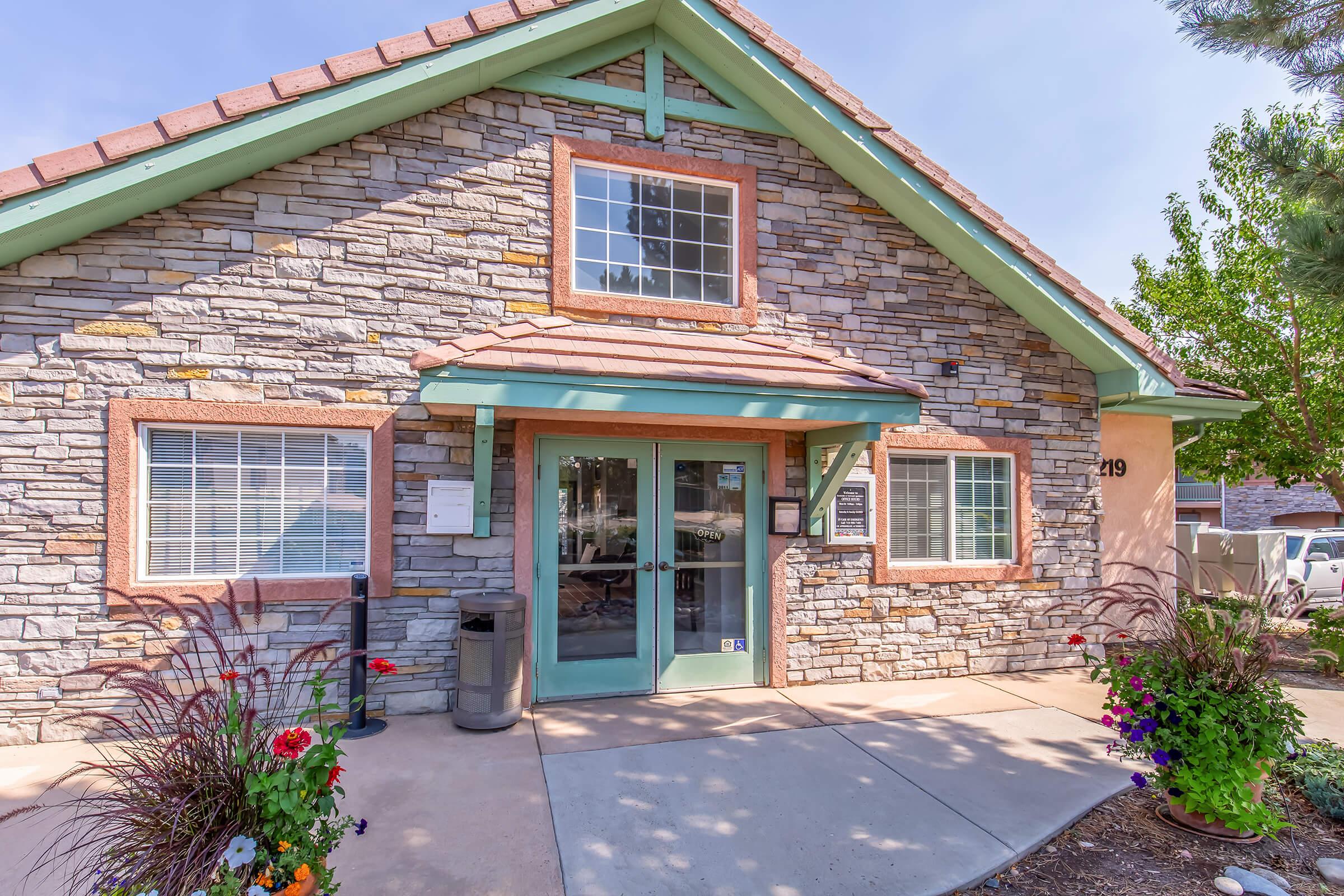 A charming stone building with a teal accent and a peaked roof. The entrance features double glass doors, flanked by large windows. Colorful flower beds with various plants enhance the entrance, complementing a well-maintained path. Lush greenery surrounds the building, creating an inviting atmosphere.