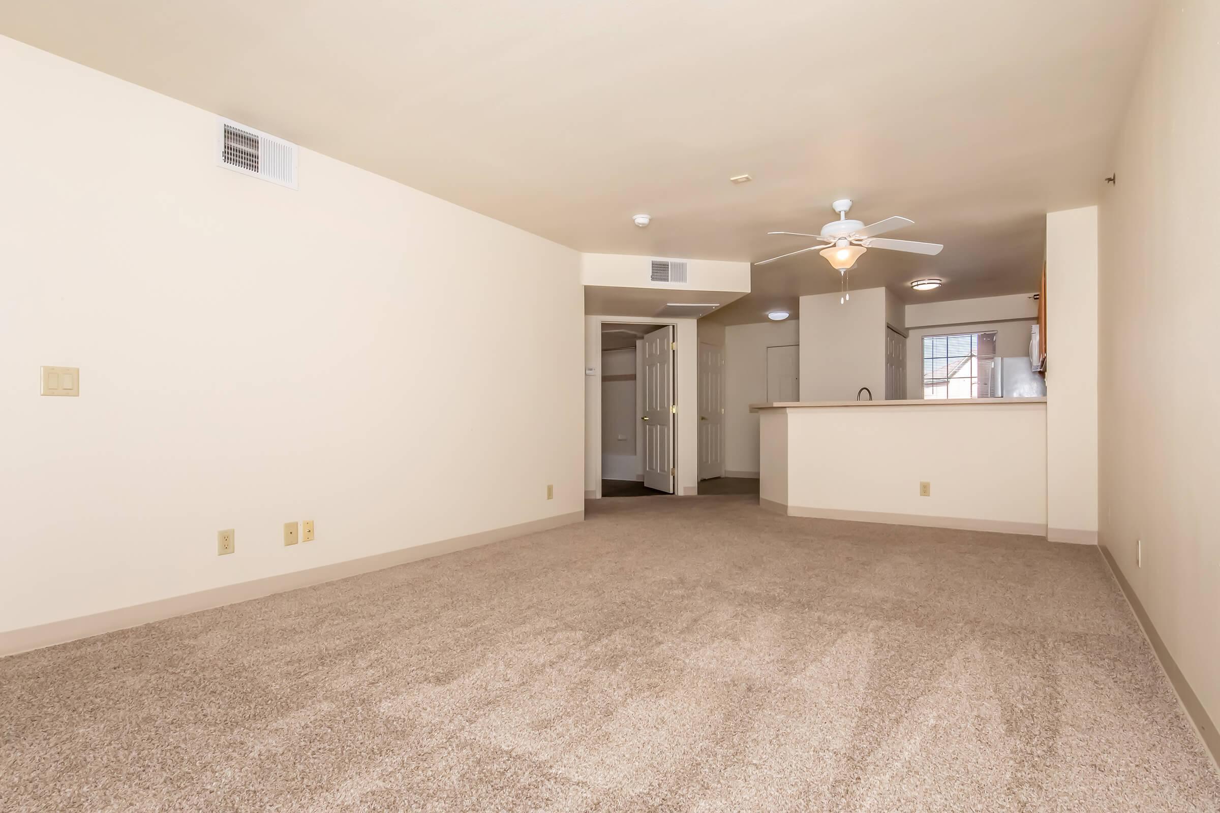 A spacious, vacant living room with light beige carpet and cream-colored walls. A ceiling fan hangs from the ceiling, and there is a small kitchenette in the background with an open design. The room is well-lit with natural light coming from a window, creating a bright and airy atmosphere.