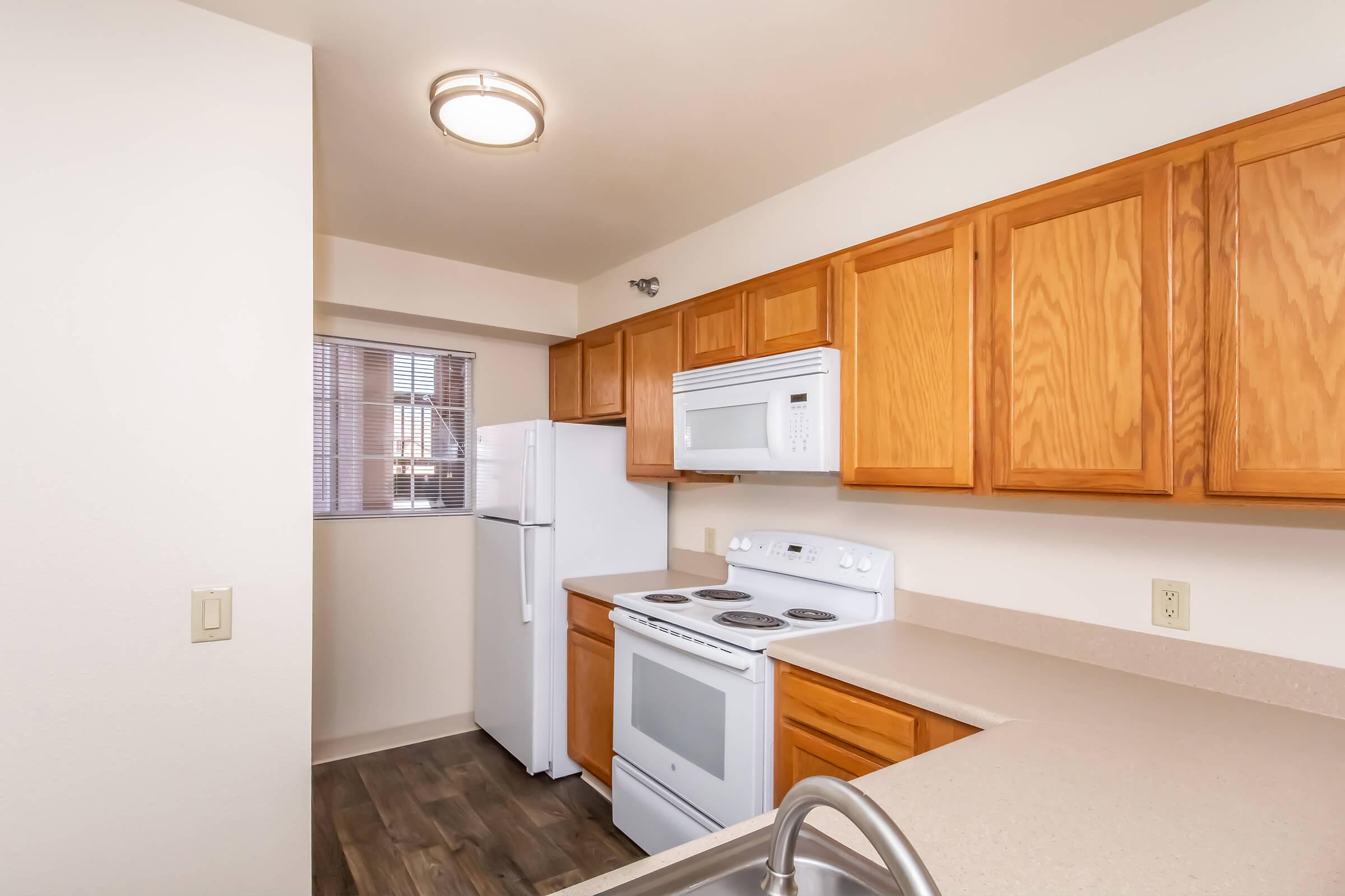 A modern kitchen featuring wooden cabinets, a white oven, a microwave, and a refrigerator. The countertop is light-colored, and there's a window allowing natural light. The flooring appears to be laminate. Overall, the kitchen has a clean and inviting appearance.