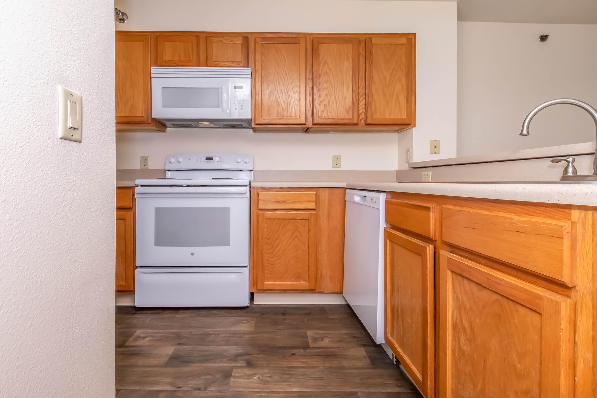 Modern kitchen featuring wooden cabinets, a countertop, a white microwave above a white stove, and a dishwasher. The flooring is dark wood, and the walls are light-colored, creating a bright and inviting space.