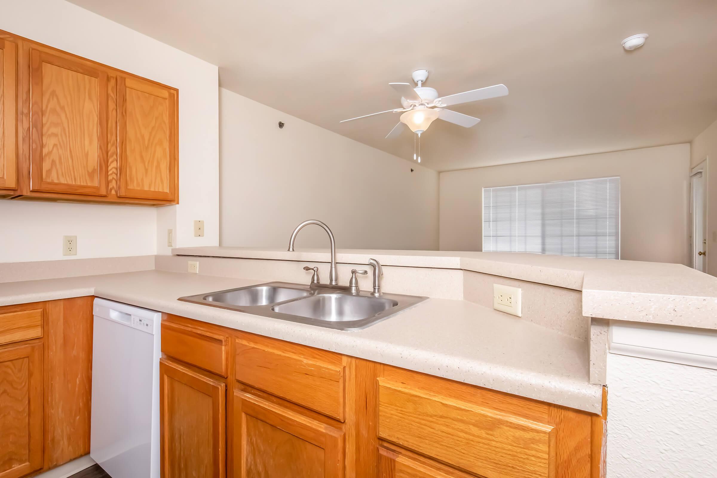 A spacious kitchen featuring wooden cabinets, a double sink, and a dishwasher, with a light-colored countertop. A ceiling fan is visible above, and there's a bright living area in the background with a large window and vertical blinds. The overall design is light and open.