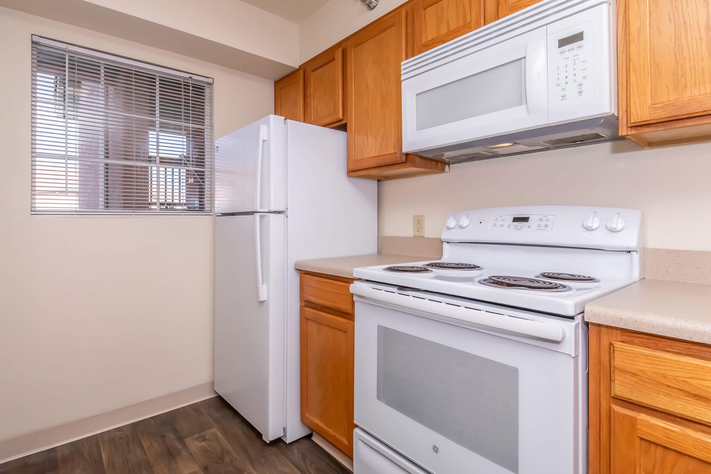 A modern kitchen featuring light wooden cabinets, a white refrigerator, a white stove and oven, and a built-in microwave. There is a window with blinds allowing natural light, and a neutral-colored countertop complements the appliances. The floor is a dark wood laminate.