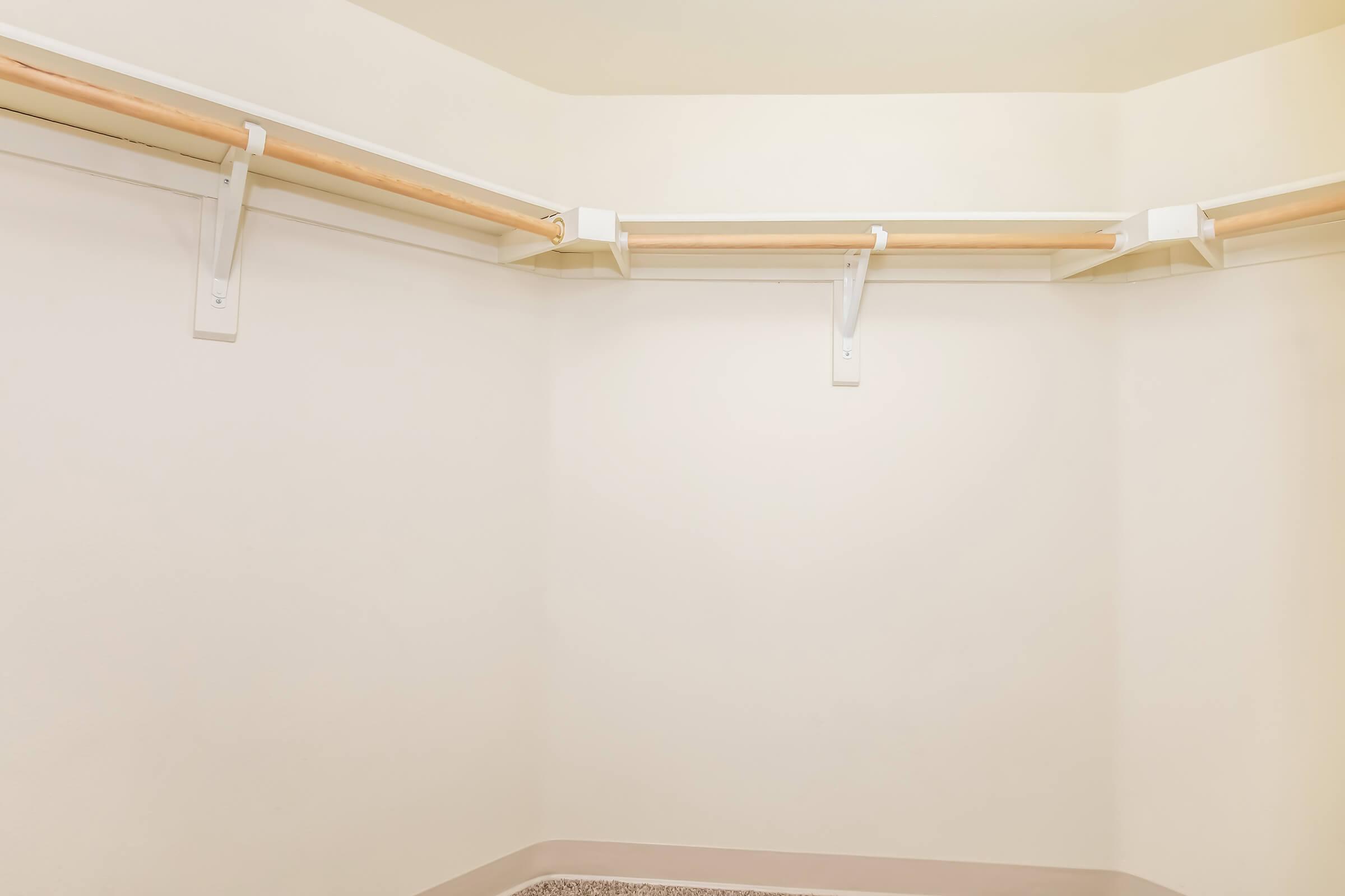 Empty corner closet with light-colored walls and two wooden clothes rods mounted on white brackets. The floor is covered with a light-colored carpet, creating a clean and minimalistic appearance.